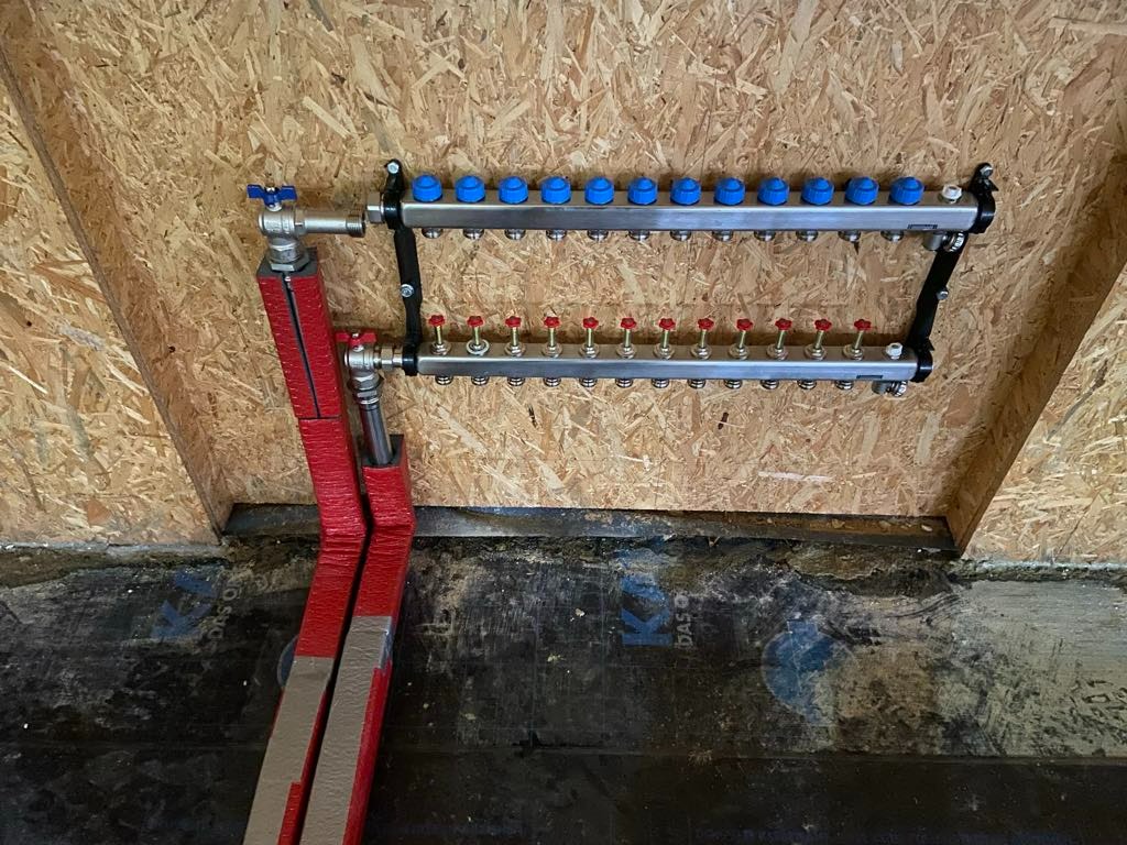 Rozdzielacz do instalacji grzewczej z niebieskimi i czerwonymi zaworami zamontowany na drewnianej ścianie, rury w czerwonej izolacji.