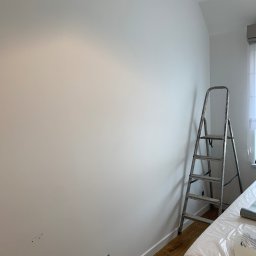 Katarzyna Trofiniak - Biała ściana po malowaniu, z widoczną drabiną i zabezpieczonymi meblami folią. Gniazdko elektryczne w ścianie, drewniana podłoga.