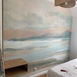 Katarzyna Trofiniak - Malowanie ściany z motywem pejzażu w pokoju, widoczna drabina, szafka i zabezpieczone łóżko. Ściana w odcieniach beżu, błękitu i szarości.