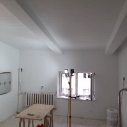 Katarzyna Trofiniak - Białe, świeżo pomalowane wnętrze pokoju z lampą budowlaną, grzejnikiem i stołem roboczym. Widoczne wykończenia sufitu i ścian po remoncie.