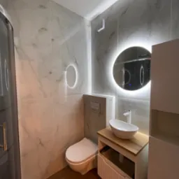 Nowoczesna łazienka z marmurowymi płytkami, okrągłym lustrem z podświetleniem LED, drewnianym blatem umywalki i kabiną prysznicową z ciemnymi ramami.