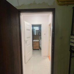 Katarzyna Trofiniak - Otwarta, biała łazienka z drewnianą szafką pod umywalką, widoczna przez futrynę drzwi. W tle toaleta i lustro. Świeże wykończenie, jasne płytki podłogowe.