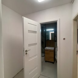 Katarzyna Trofiniak - Otwarta biała łazienkowa z drewnianą szafką pod umywalką i kabiną prysznicową, widoczna z korytarza. Minimalistyczny design, jasne oświetlenie.