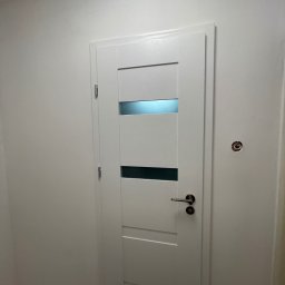 Katarzyna Trofiniak - Białe, nowoczesne drzwi wewnętrzne z przeszkleniami, zamontowane w świeżo pomalowanym na biało pomieszczeniu. Widoczny fragment podłogi i ściany z otworem po instalacji.