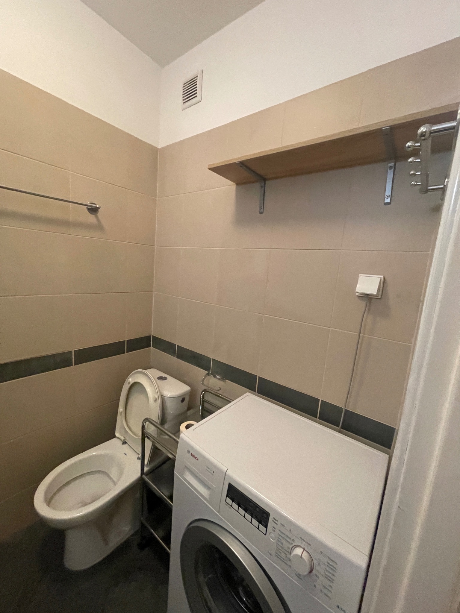Ustawna łazienka w beżowych płytkach z ciemnym pasem, z toaletą, pralką Bosch i drewnianą półką. Wykończenie w neutralnej kolorystyce.