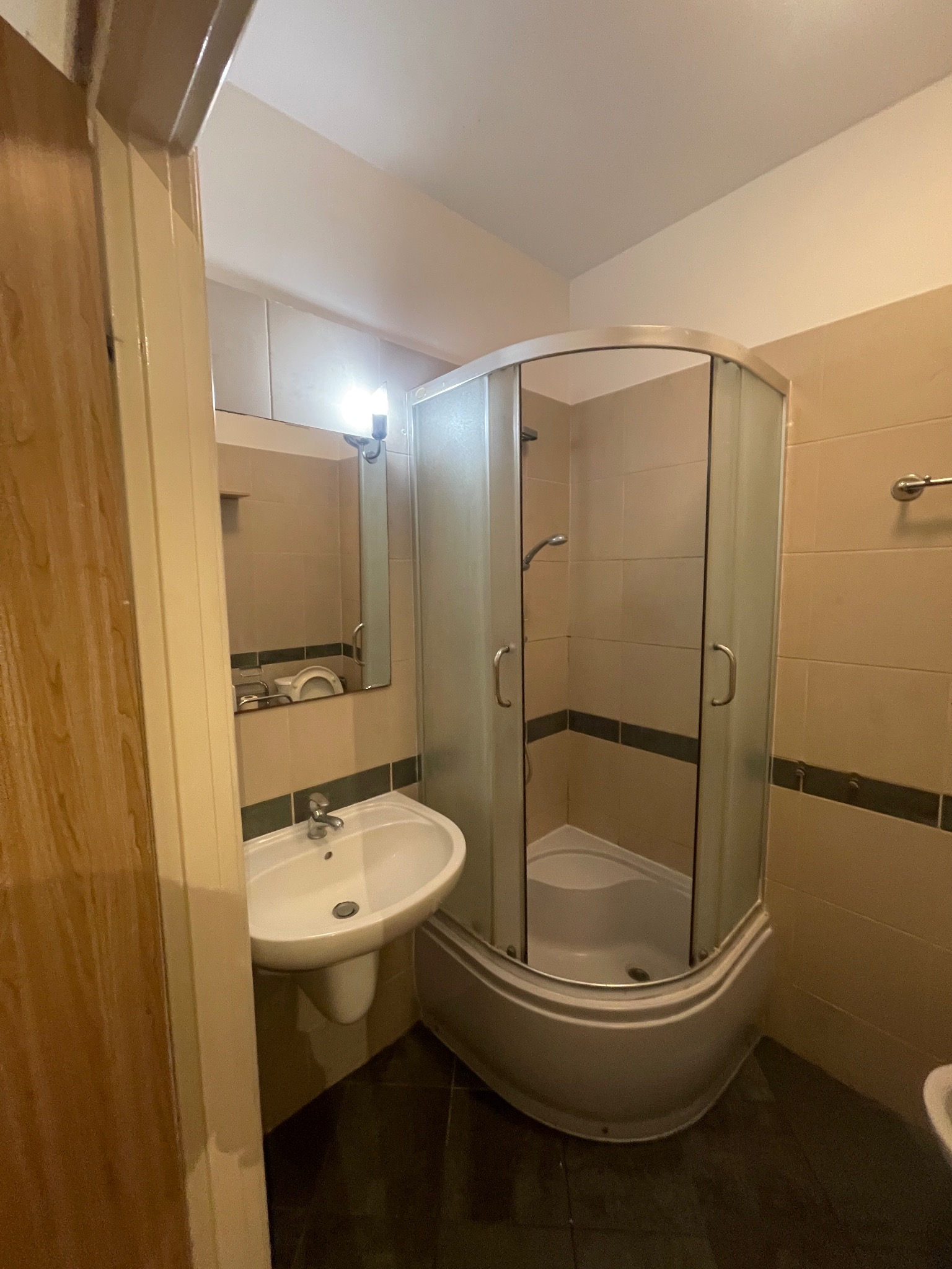 Wykończona łazienka z kabiną prysznicową narożną i umywalką na postumencie, widok zza otwartych drzwi. Neutralna kolorystyka płytek i armatury.