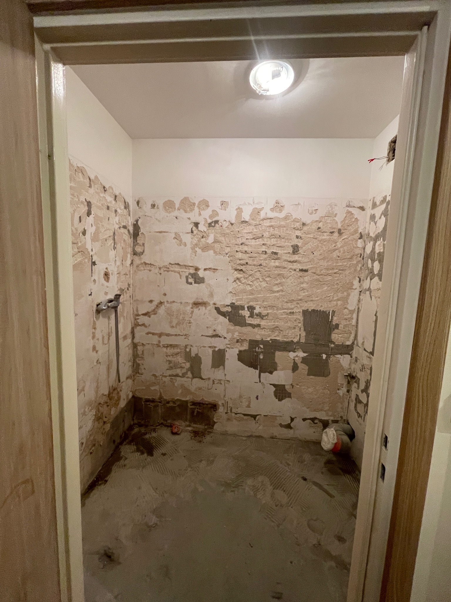 Wnętrze łazienki w trakcie demontażu płytek, widoczne ślady po kleju i fragment instalacji wodnej. Odsłonięte ściany z resztkami starej glazury i przygotowane podłoże.