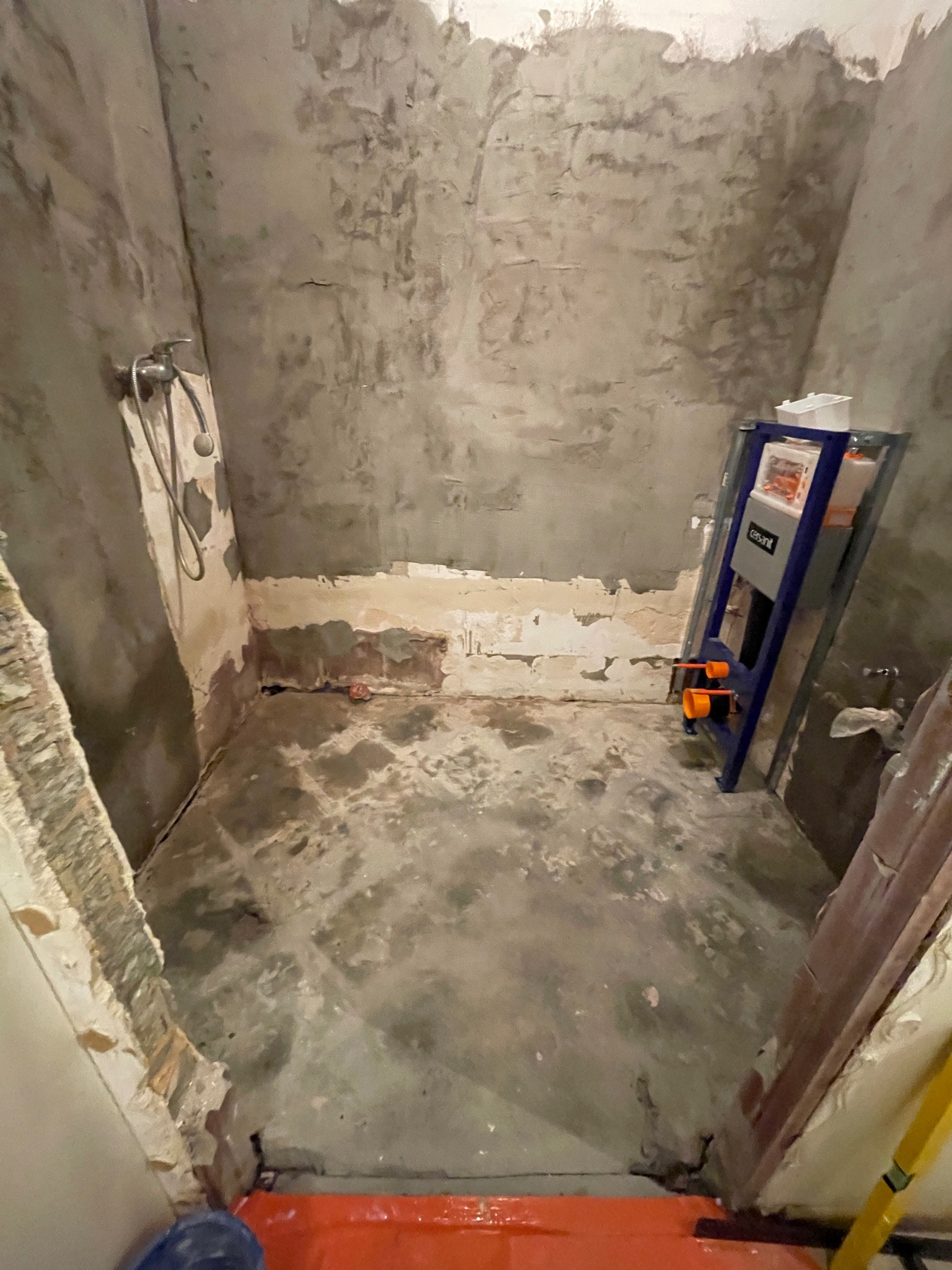 Surowy stan łazienki w trakcie remontu: widoczne ściany z resztkami tynku, zamontowany stelaż podtynkowy WC, fragment instalacji prysznicowej i poziomica.