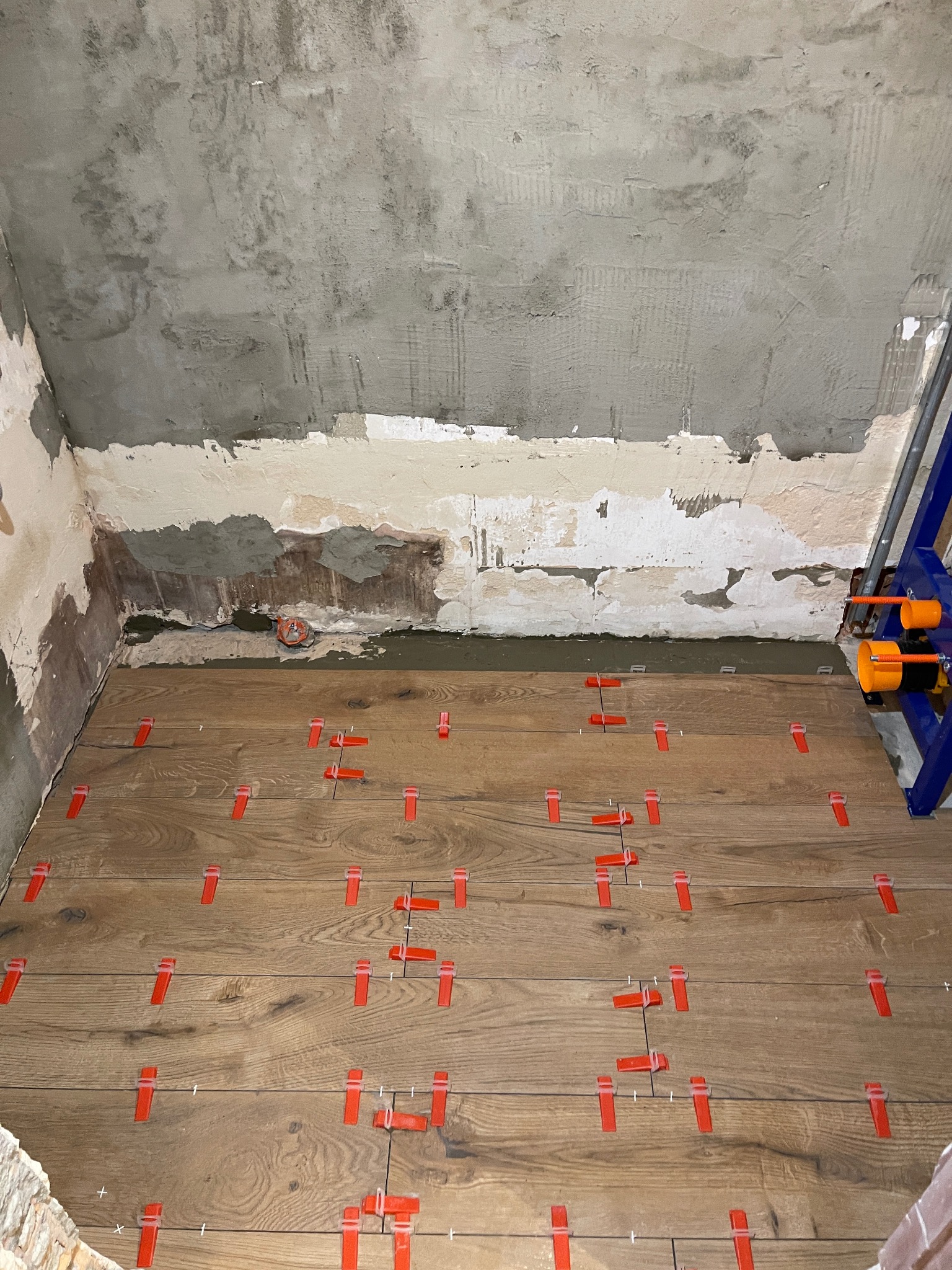 Układanie drewnopodobnych płytek na podłodze z użyciem czerwonych klipsów poziomujących. Ściany surowe, przygotowane do dalszych prac wykończeniowych.