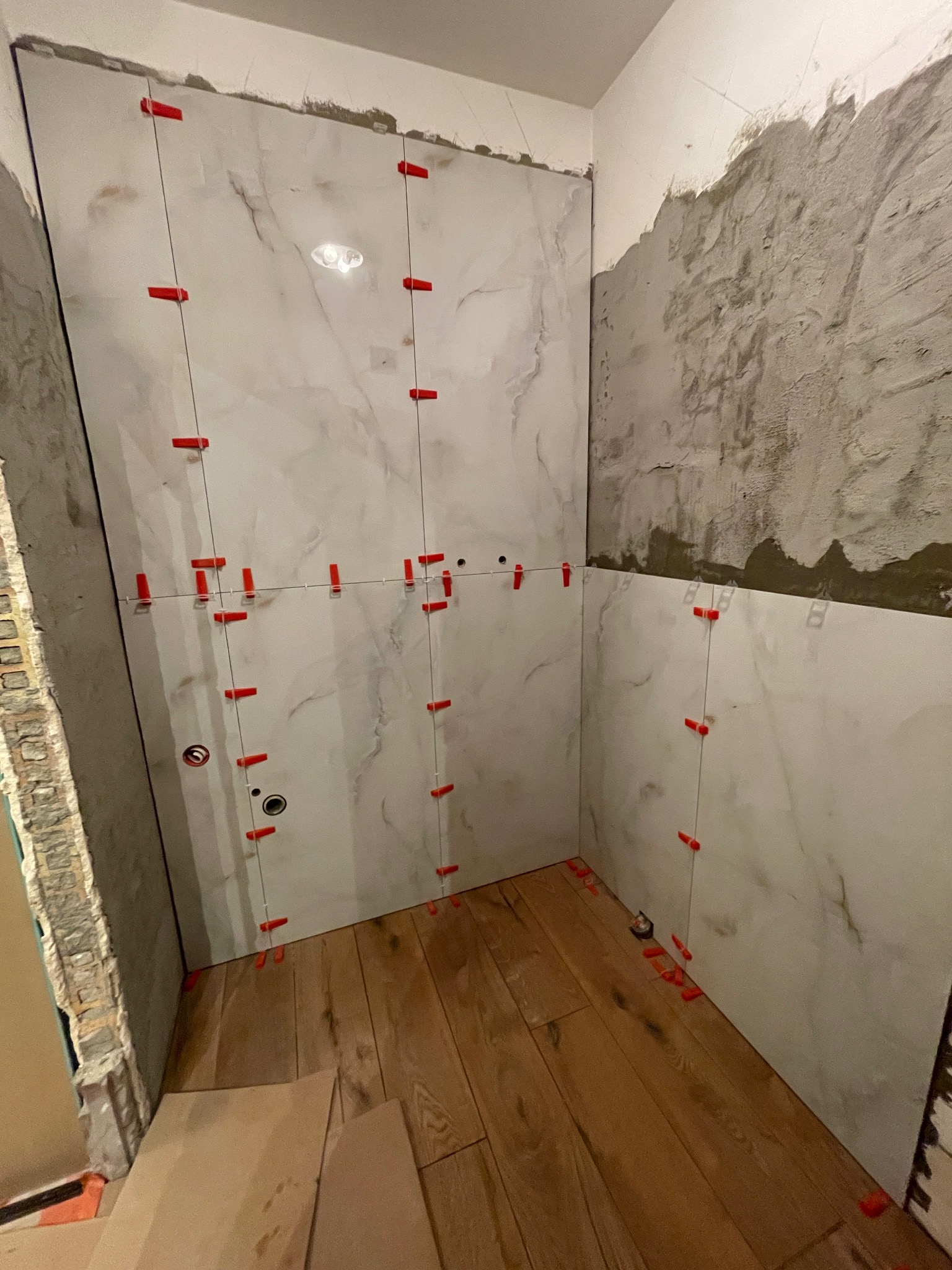 Układanie płytek marmurowych w łazience, widoczne czerwone klipsy poziomujące, drewnopodobna podłoga, surowy tynk na ścianie, etap prac wykończeniowych.