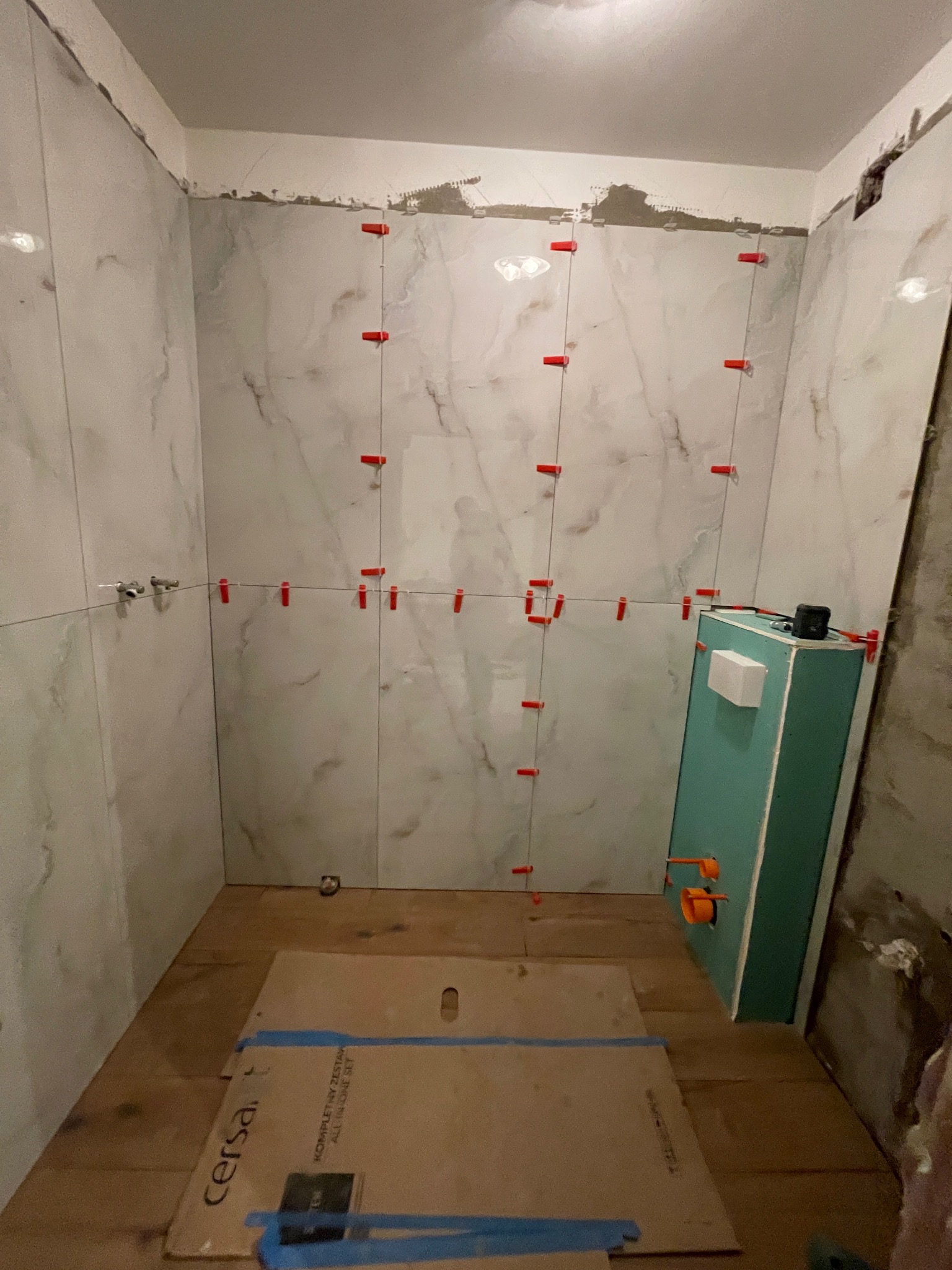 Łazienka w trakcie remontu: białe płytki imitujące marmur na ścianach, czerwone krzyżyki dystansowe, zabudowa WC w kolorze turkusowym, kartony na podłodze.
