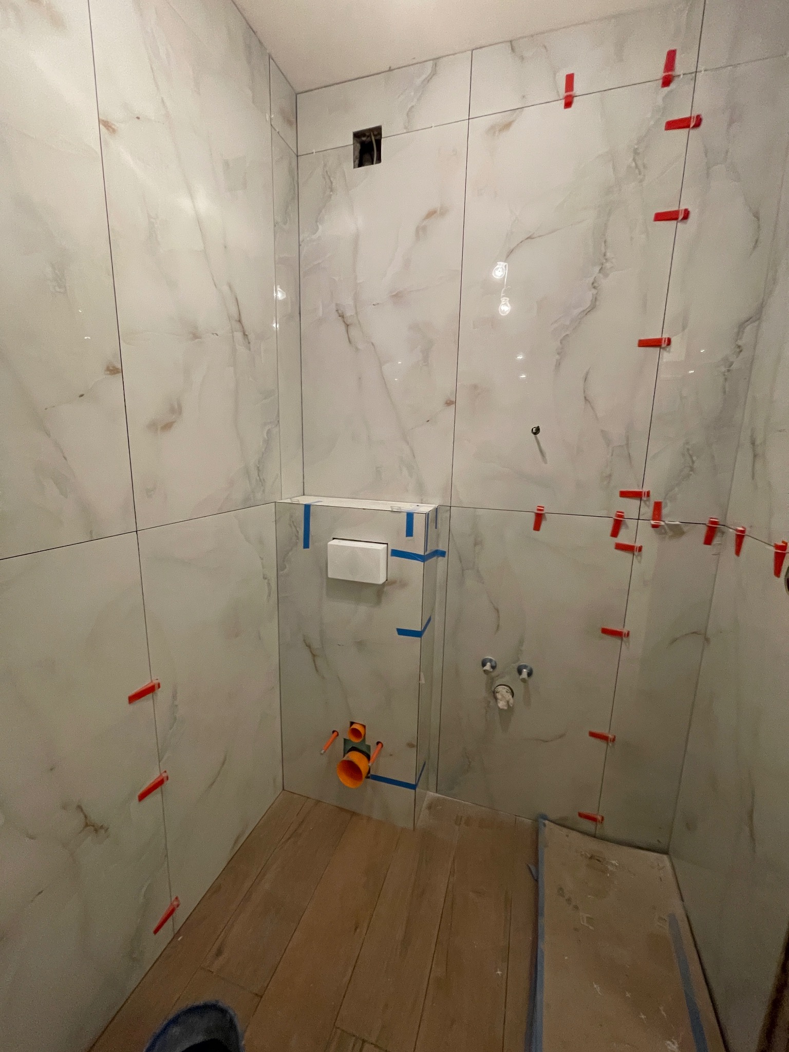 Łazienka w trakcie wykończenia: białe płytki imitujące marmur, czerwone krzyżyki dystansowe, widoczne rury i stelaż podtynkowy WC, drewnopodobna podłoga.