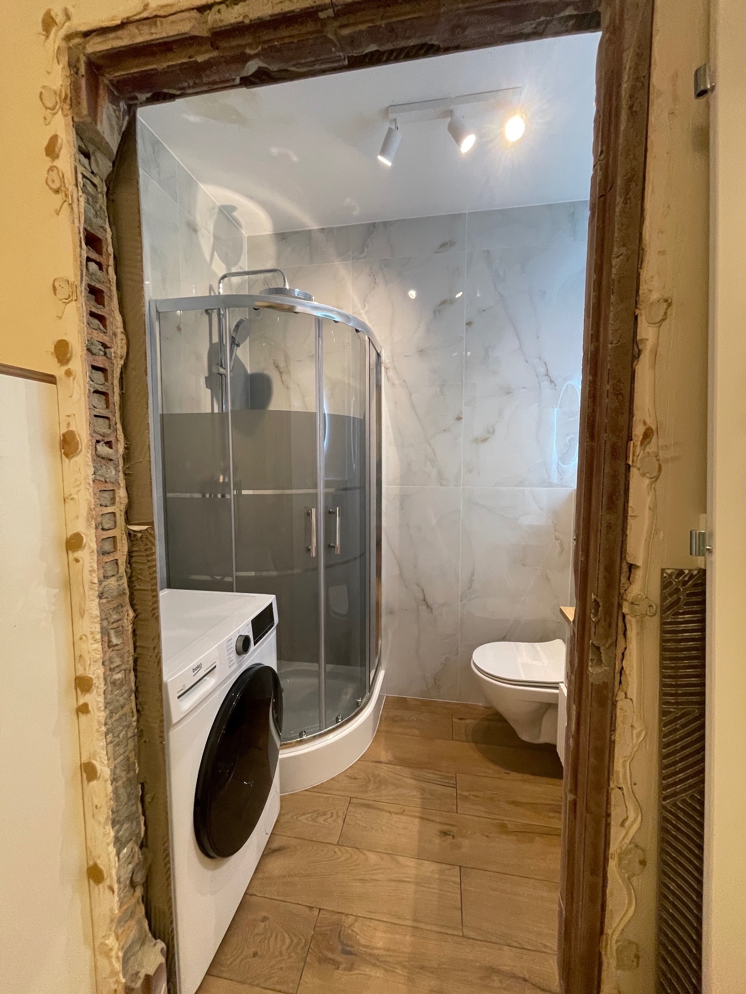 Wykończona łazienka z kabiną prysznicową, toaletą i pralką, widziana przez surowe, częściowo obrobione ościeże drzwi.