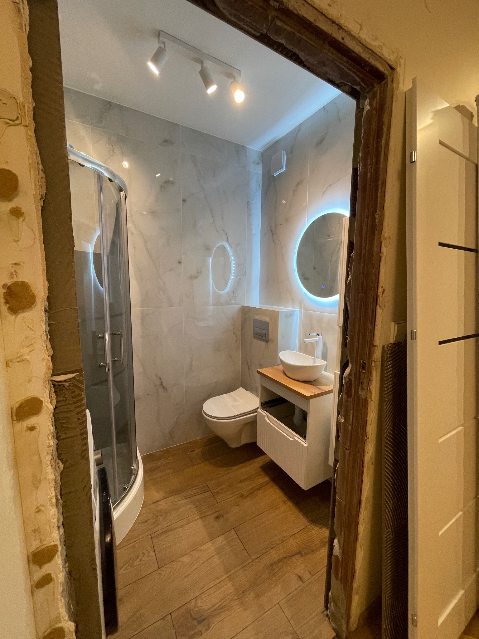Nowoczesna łazienka z kabiną prysznicową, umywalką na drewnianym blacie i okrągłym lustrem z podświetleniem LED, widziana przez surową futrynę w trakcie remontu.