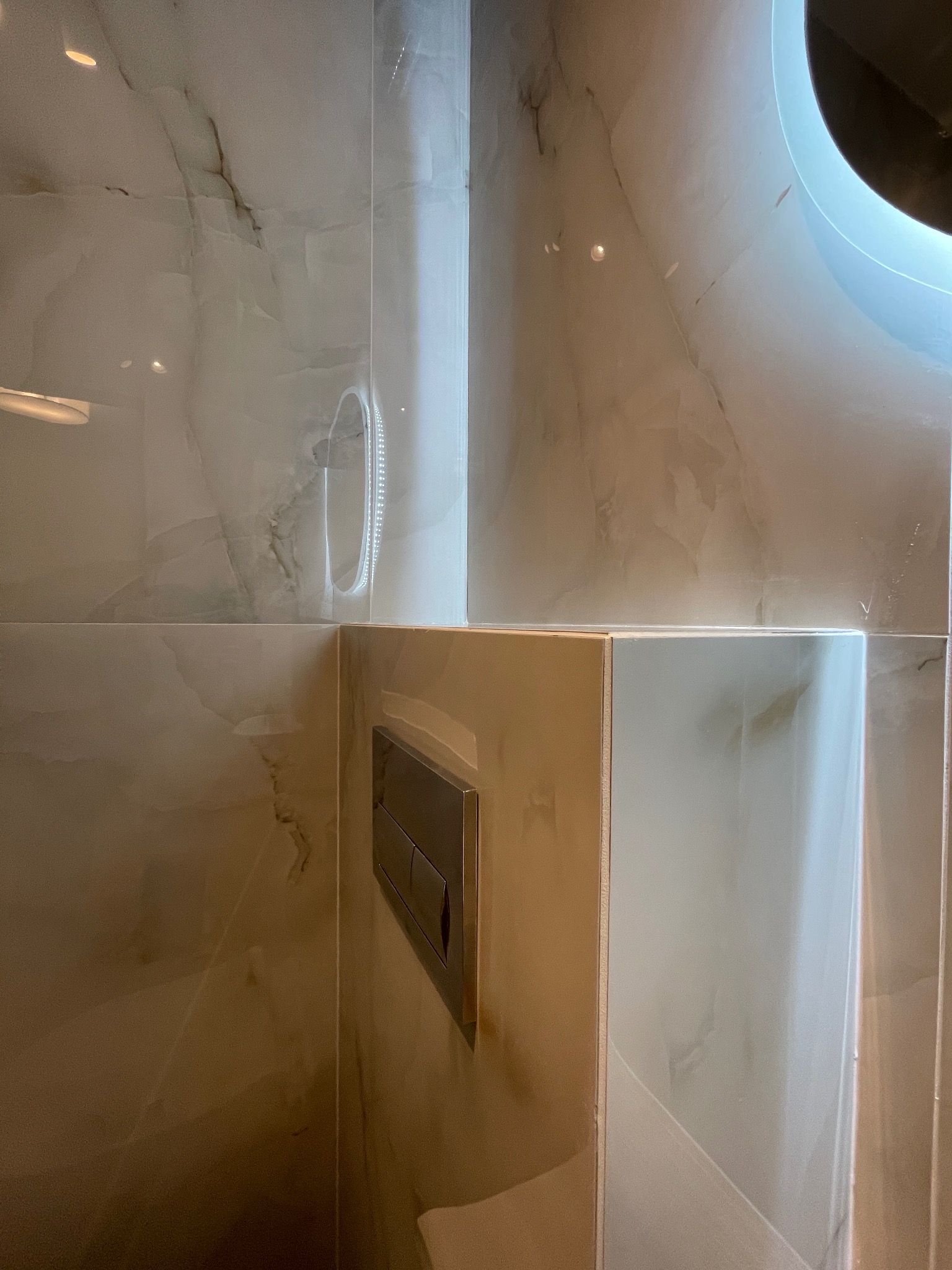 Elegancka łazienka z marmurowymi płytkami, widoczny panel sterujący spłuczką i fragment oświetlonego lustra. Minimalistyczny design i subtelne oświetlenie.