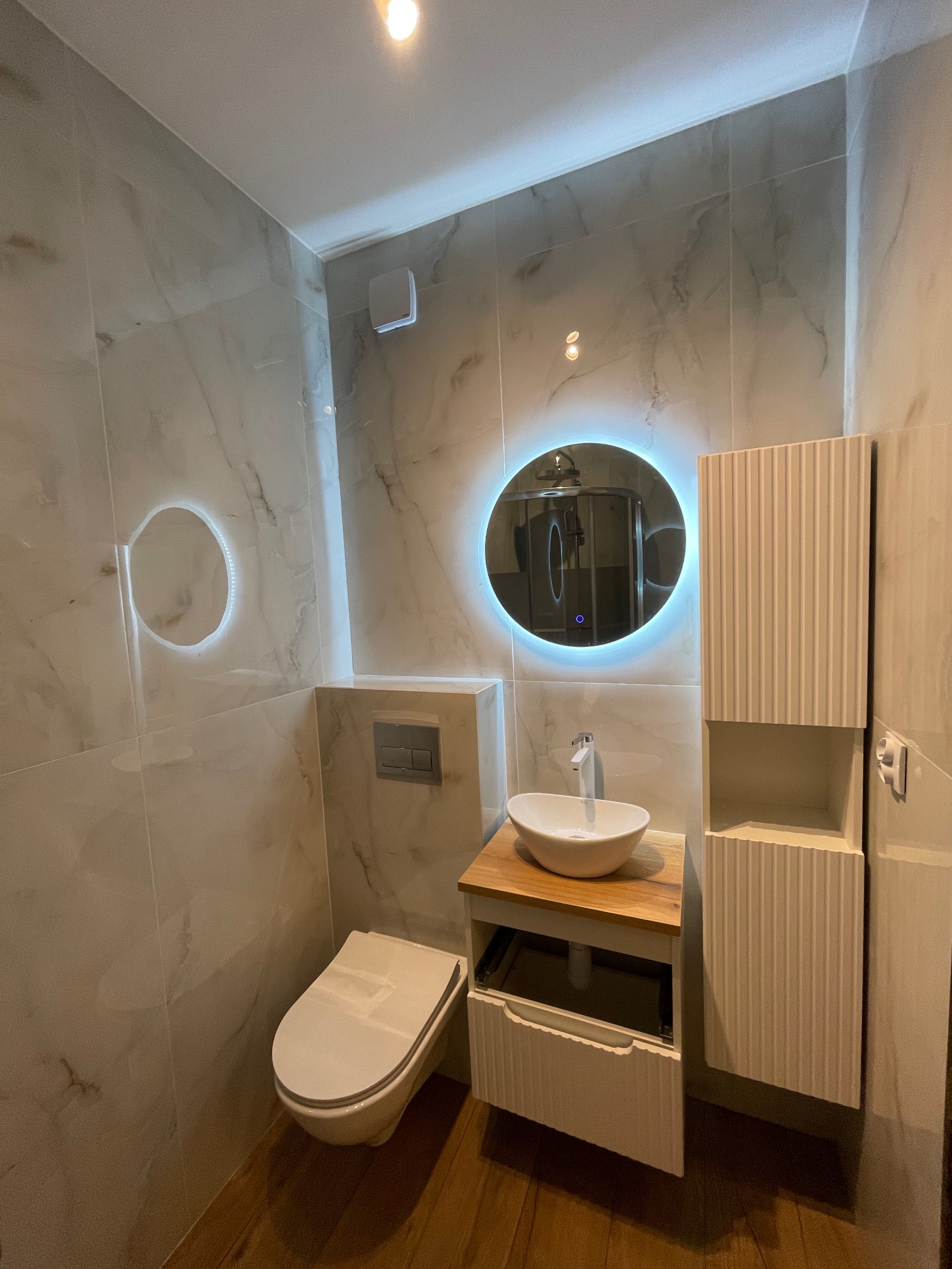 Nowoczesna łazienka z marmurowymi płytkami, okrągłym lustrem z podświetleniem LED, wiszącą toaletą i szafkami z ryflowanymi frontami. Drewniany blat pod umywalką.