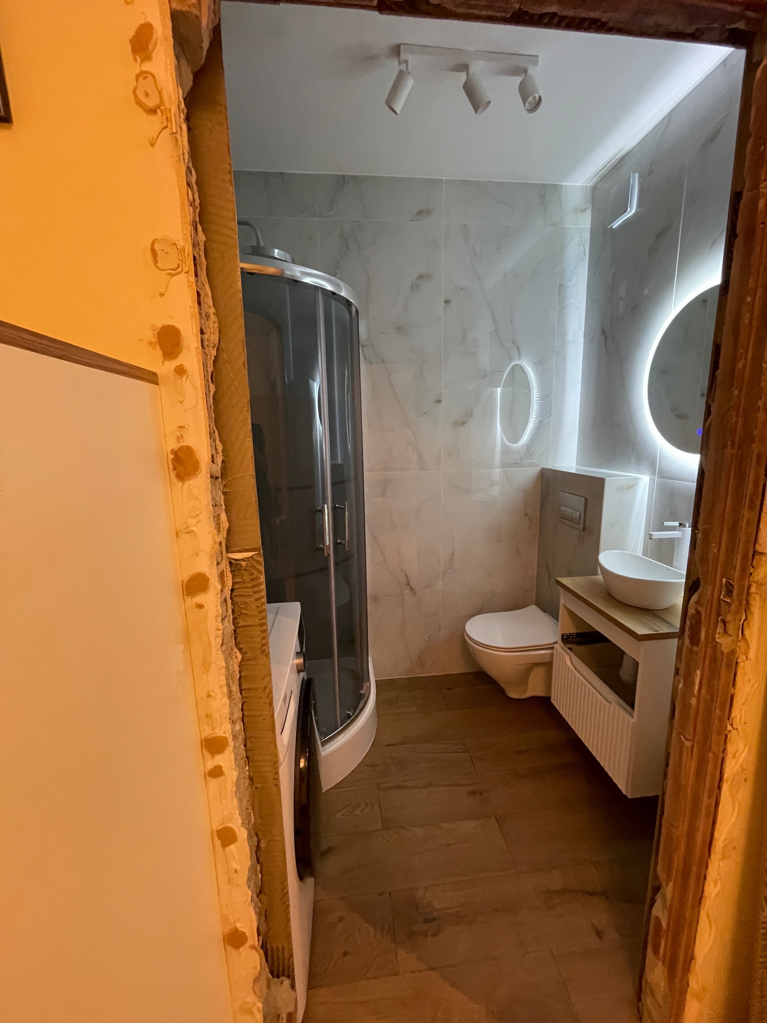 Nowoczesna łazienka z kabiną prysznicową, umywalką na szafce i podświetlanym lustrem, widziana przez surowy otwór drzwiowy w trakcie remontu. Wykończenie w bieli i szarości.