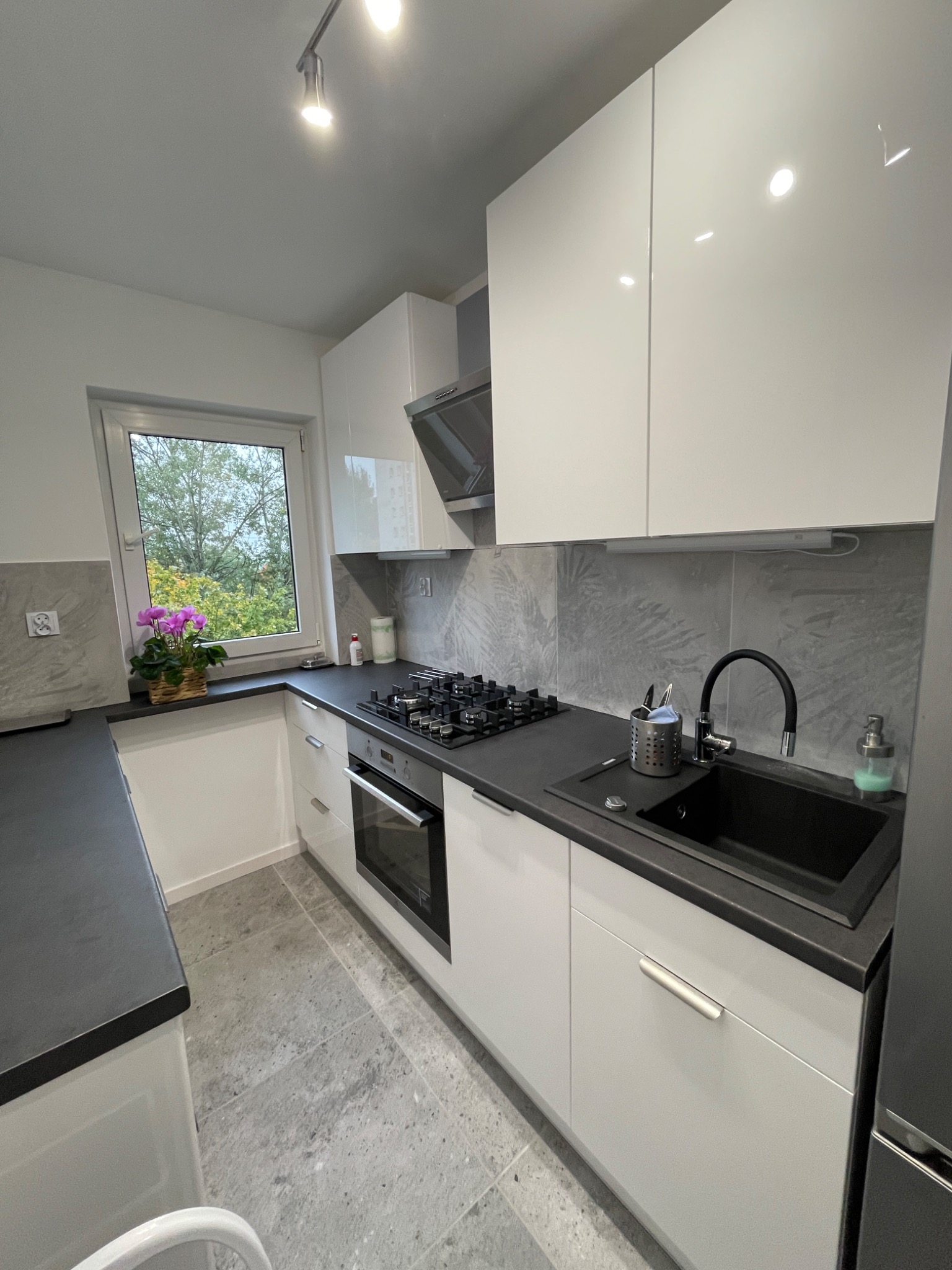 Nowoczesna kuchnia z białymi szafkami, czarnym blatem i zlewozmywakiem. Na blacie kwiaty w doniczce przy oknie. Szare płytki na ścianie i podłodze.