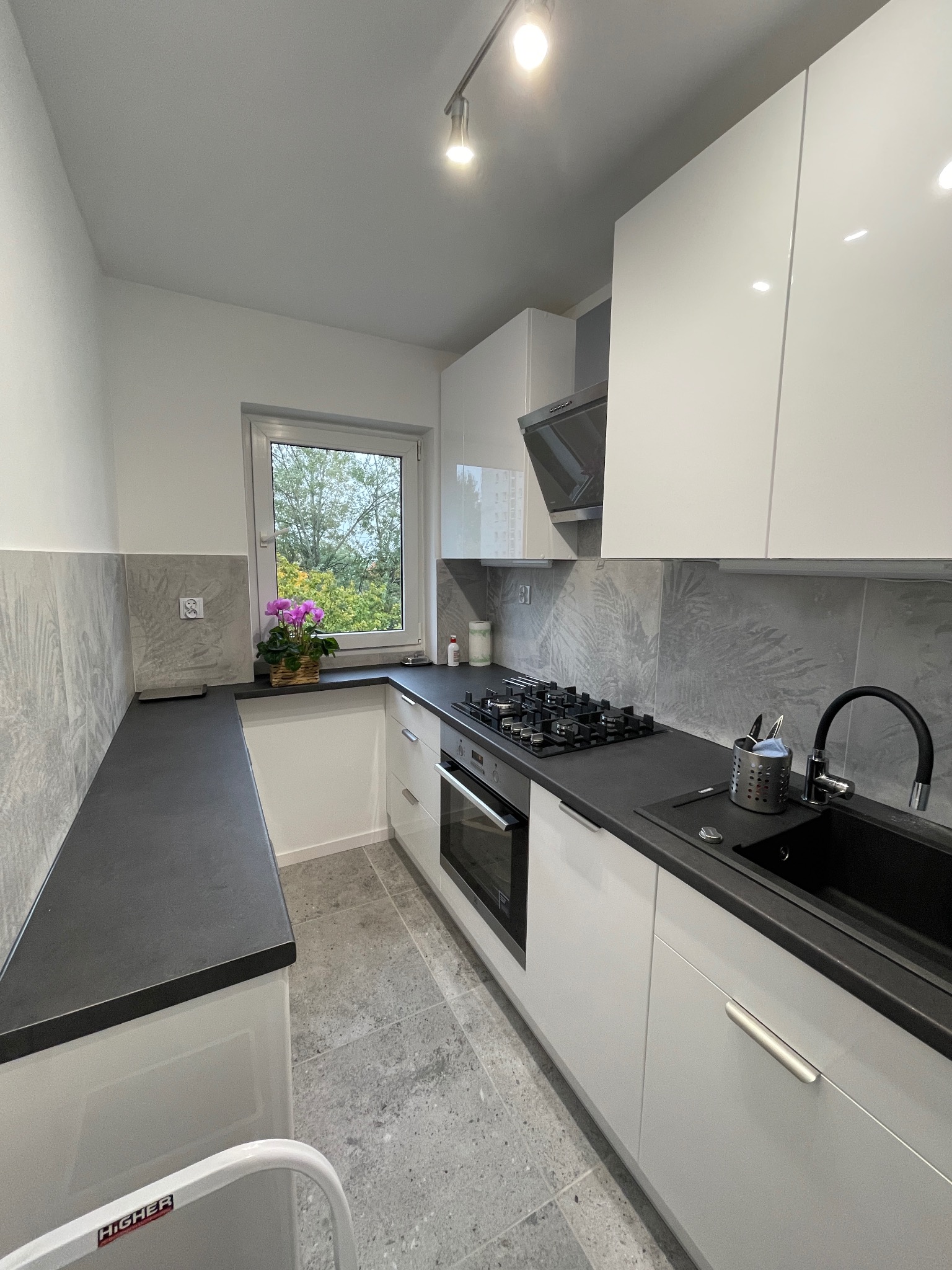 Nowoczesna kuchnia z białymi, błyszczącymi szafkami, czarnym blatem i płytkami z motywem liści. Widok na okno z kwiatami i fragment drabiny.