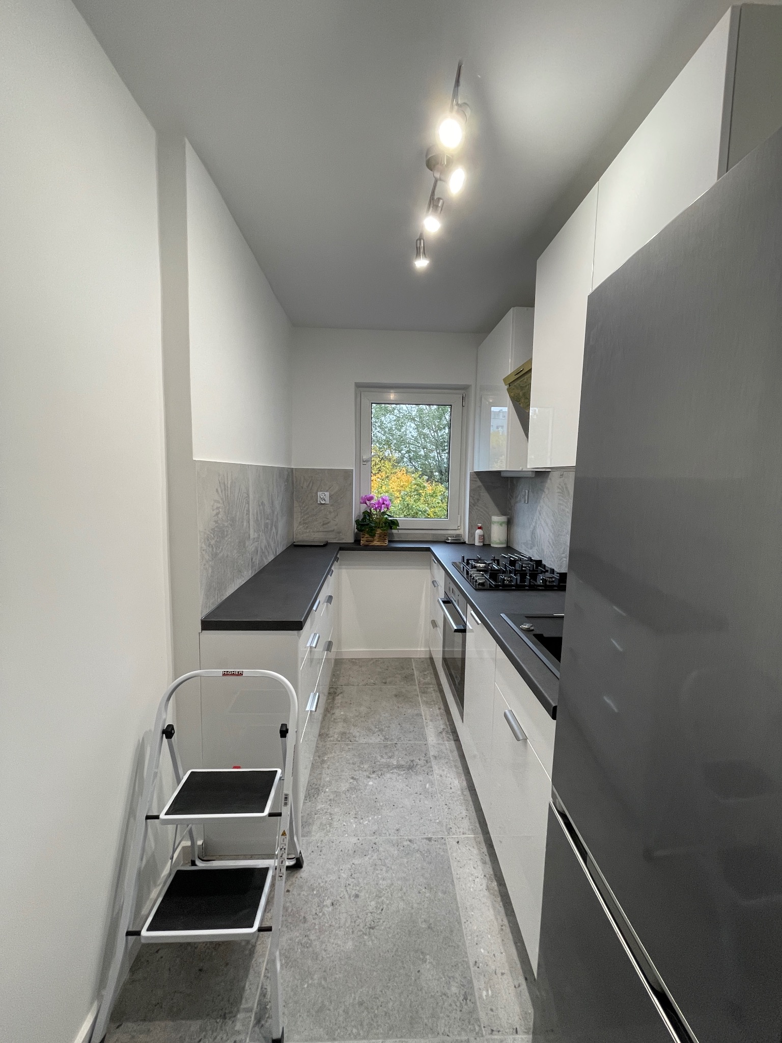 Wąska, jasna kuchnia z szarymi akcentami: białe szafki z czarnym blatem, szara podłoga i dekoracyjny panel nad blatem. Widok na zewnątrz przez okno.