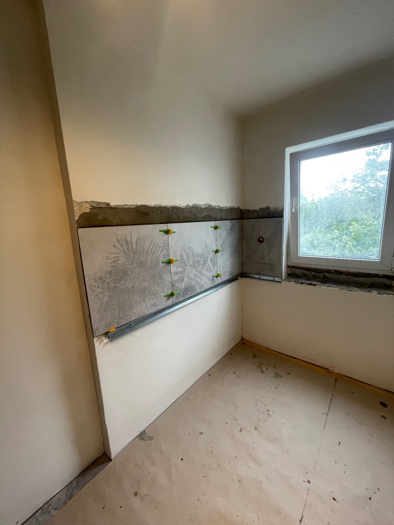 Wnętrze w trakcie prac glazurniczych: szare płytki z motywem roślinnym, zielone krzyżyki dystansowe, metalowe listwy, okno z widokiem na zieleń. Surowy stan wykończenia.