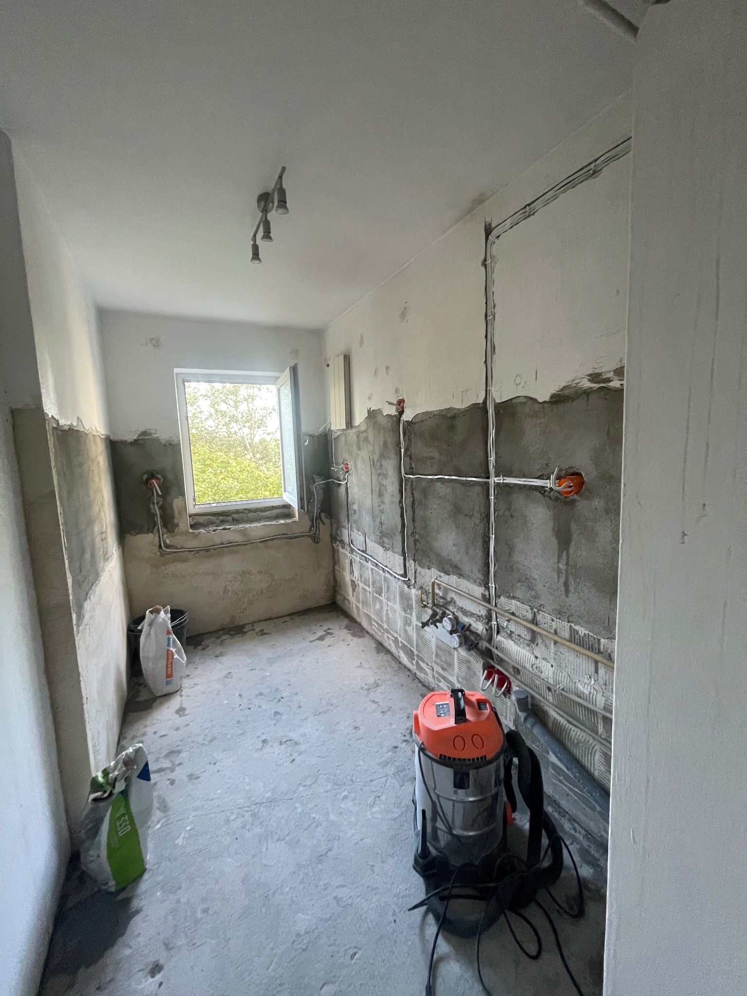 Wnętrze łazienki w trakcie prac remontowych: widoczne instalacje elektryczne, częściowo położone gładzie, worki z zaprawą i odkurzacz przemysłowy. Okno z widokiem na zieleń.