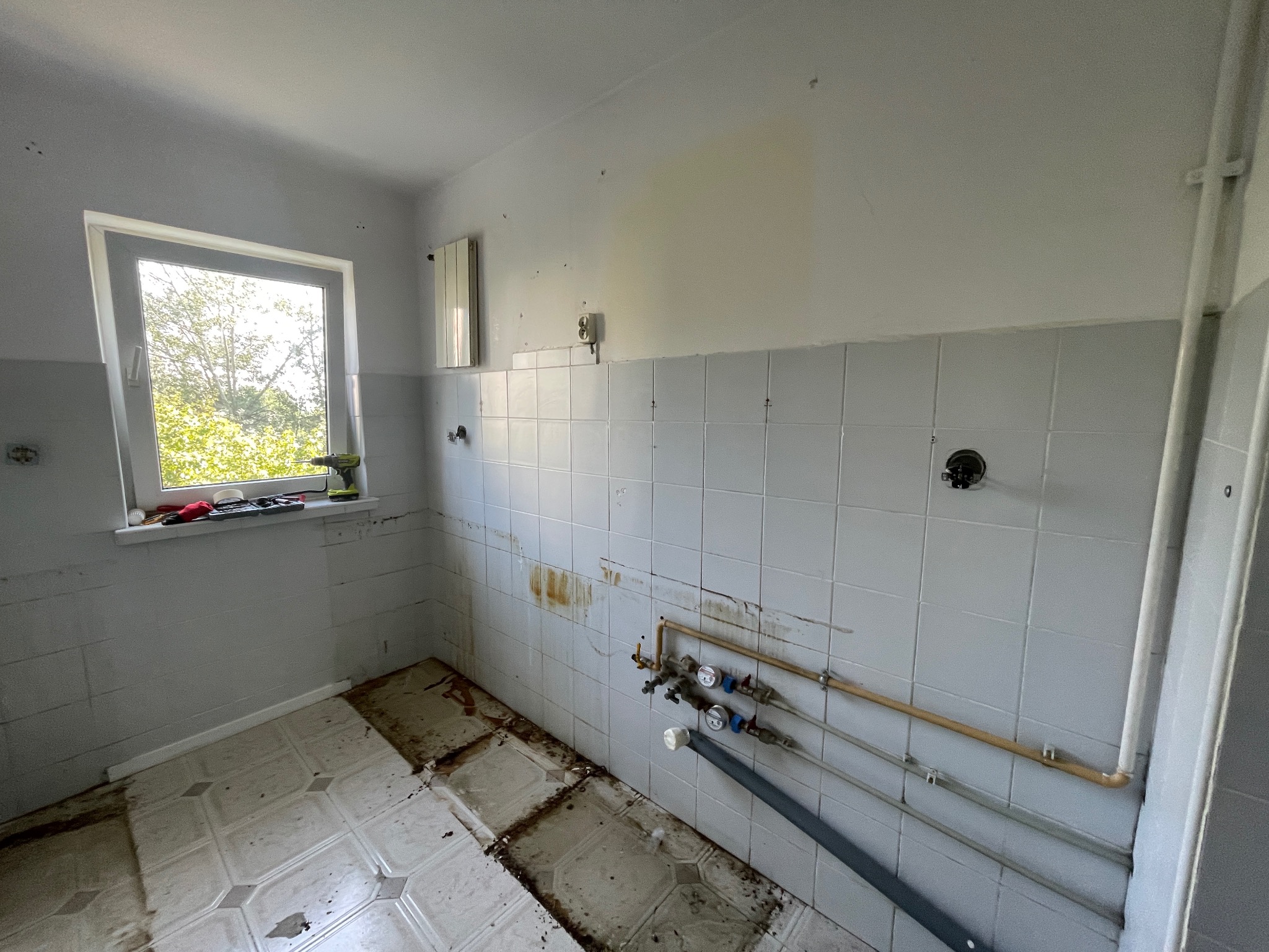 Łazienka w trakcie remontu: częściowo zerwane płytki, odsłonięte rury, narzędzia na parapecie. Widoczne ślady po starych instalacjach i przygotowanie do dalszych prac.