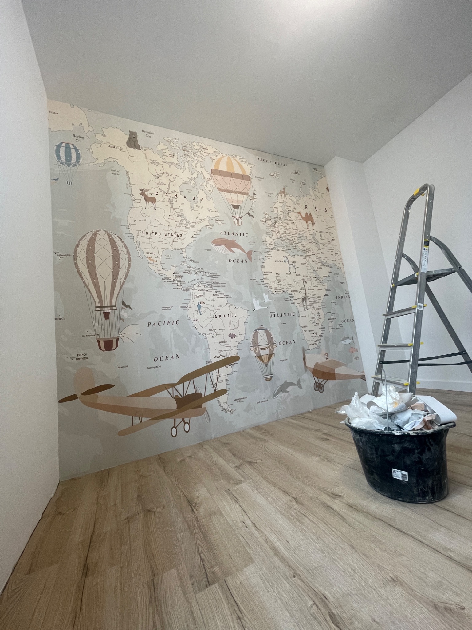 Ściana z tapetą w mapę świata z motywem podróżniczym, drabina i wiadro z odpadami na podłodze z paneli. Widok na fragment pokoju po tapetowaniu.