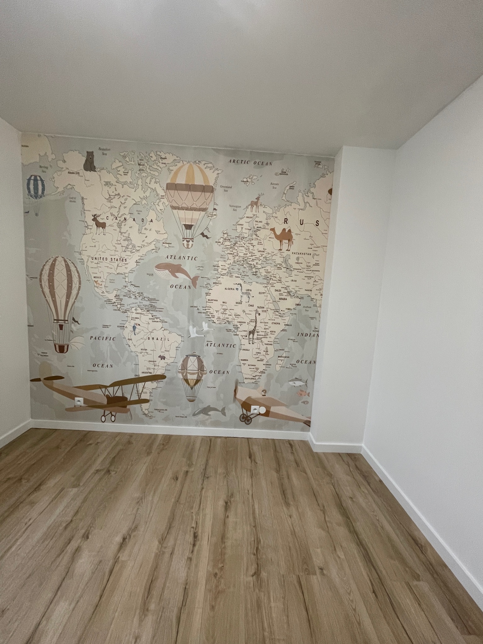 Pokój dziecięcy z drewnianą podłogą i tapetą w mapę świata z balonami i samolotami. Jasne ściany, minimalistyczny wystrój. Ujęcie z niskiego kąta, podkreślające przestrzeń.