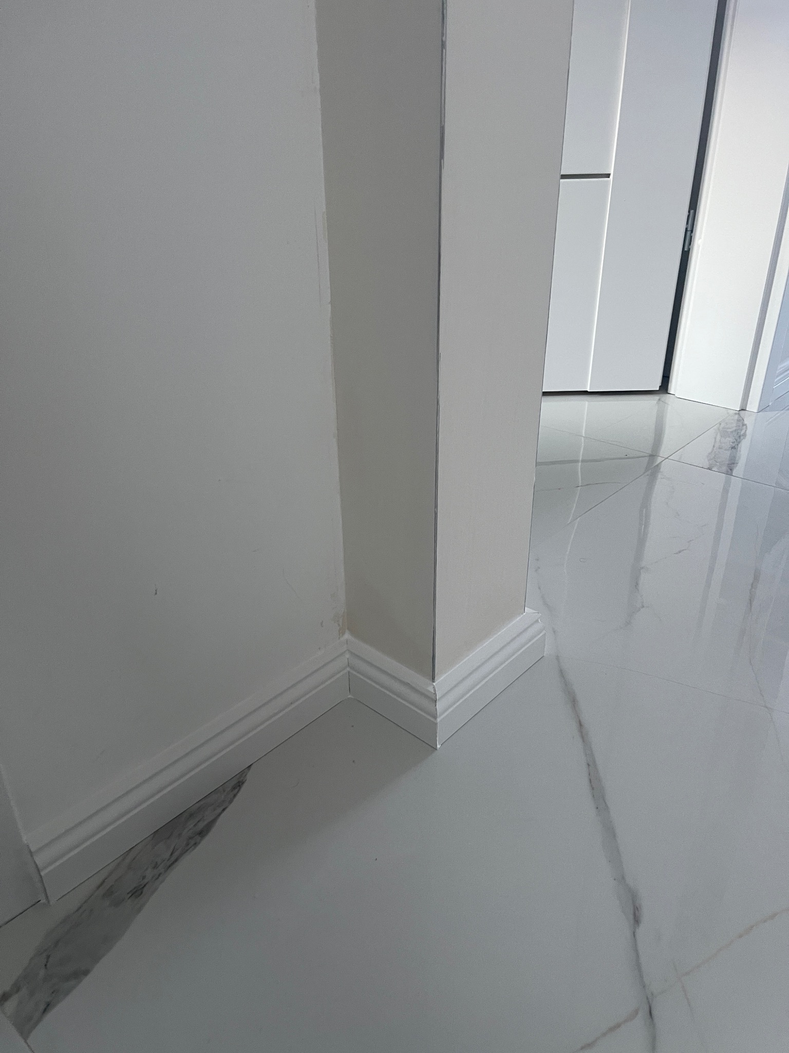 Białe listwy przypodłogowe i marmuropodobna podłoga w jasnym wnętrzu. Widoczny fragment ściany i drzwi. Minimalistyczny design.
