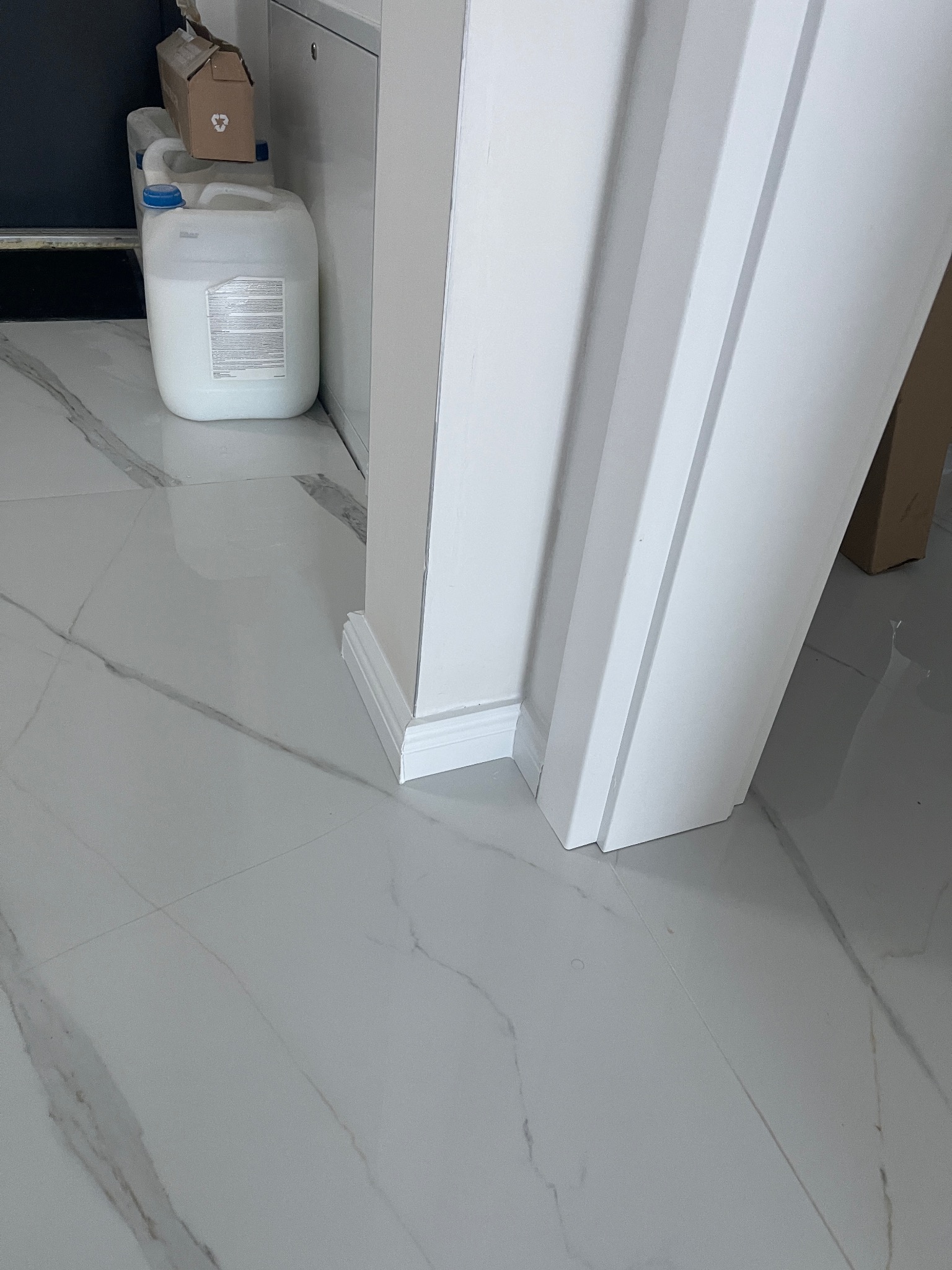 Białe listwy przypodłogowe przy ścianie i marmurowe płytki podłogowe z szarymi żyłkami. W tle pojemnik z płynem i karton. Detal wykończenia wnętrza.