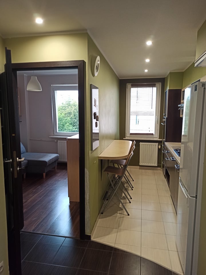 Wnętrze mieszkania po remoncie: widok z salonu na kuchnię z rozkładanym stołem i krzesłami, ciemna podłoga w salonie kontrastuje z jasną w kuchni.