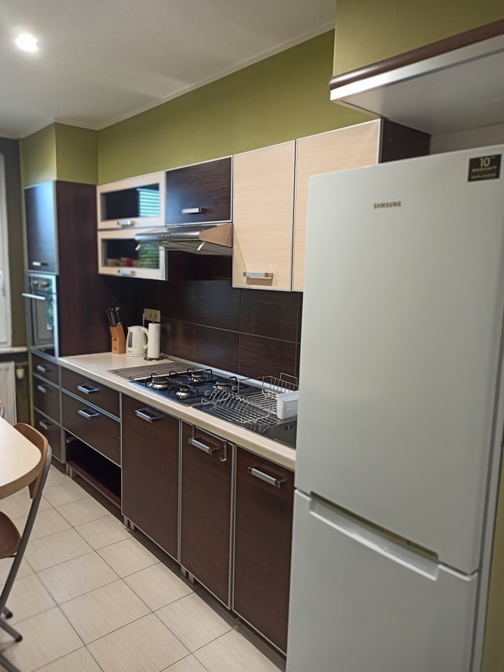 Nowoczesna kuchnia z brązowymi szafkami, białym blatem i lodówką Samsung. Widoczne płytki na ścianie i podłodze. Oświetlenie sufitowe.