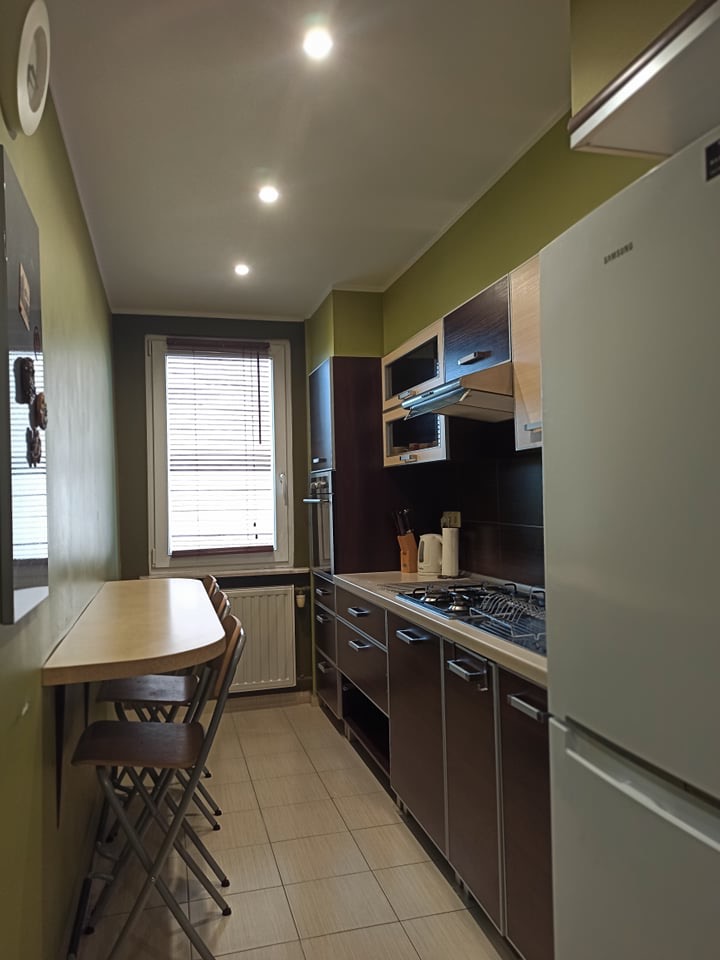 Wnętrze kuchni z brązowymi szafkami, jasnym blatem i składanym stołem z krzesłami. Widoczne okno z żaluzjami i biała lodówka. Ściany w odcieniach zieleni.