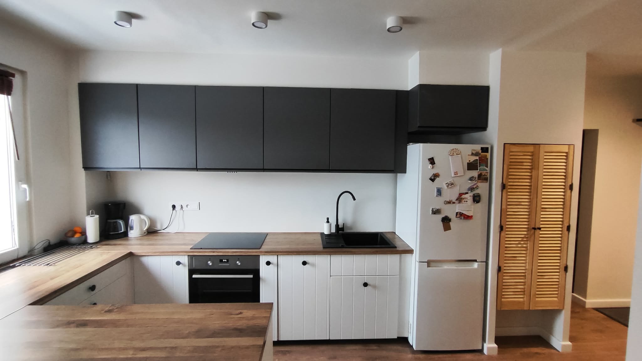 Nowoczesna kuchnia po remoncie: białe szafki z frezowanymi frontami, drewniany blat, czarny zlew i płyta indukcyjna. Górne szafki grafitowe. Lodówka z magnesami.