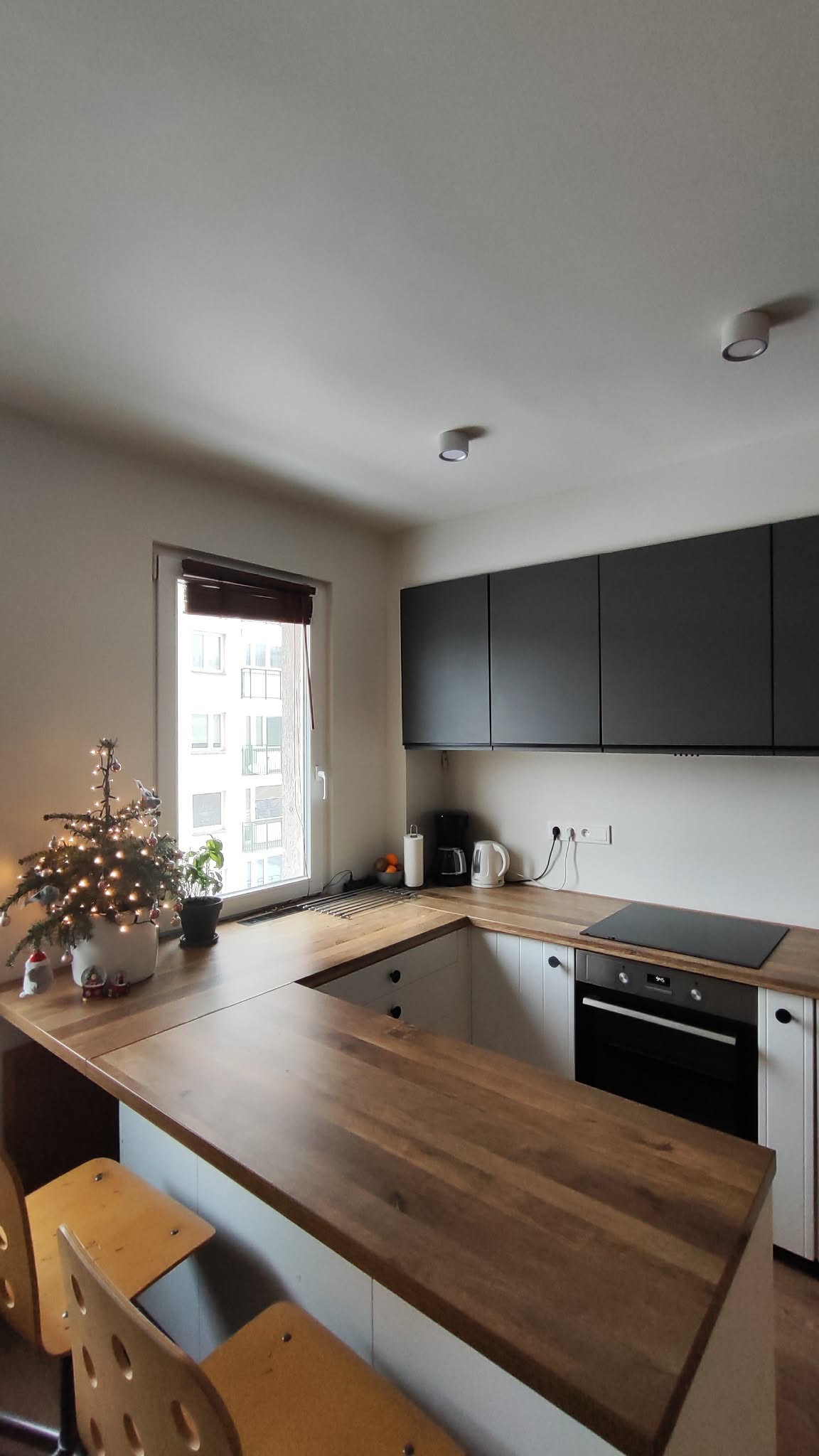 Nowoczesna kuchnia w bieli i drewnie z czarnymi szafkami wiszącymi, oświetlona światłem dziennym i punktowym, z dekoracjami świątecznymi na blacie.