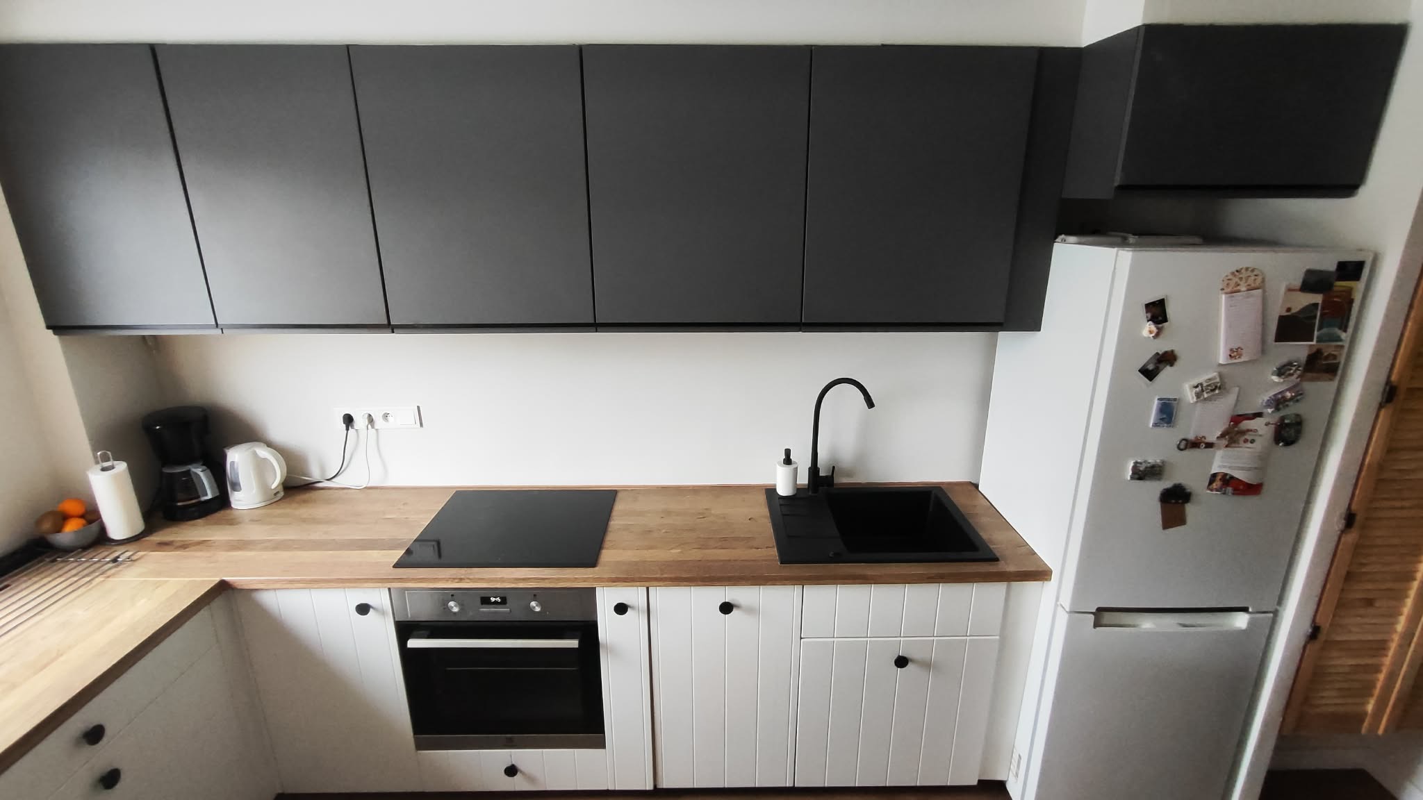Nowoczesna kuchnia z drewnianym blatem, czarnym zlewem i płytą indukcyjną. Białe szafki dolne z frezowanymi frontami i grafitowe szafki górne. Lodówka z magnesami.