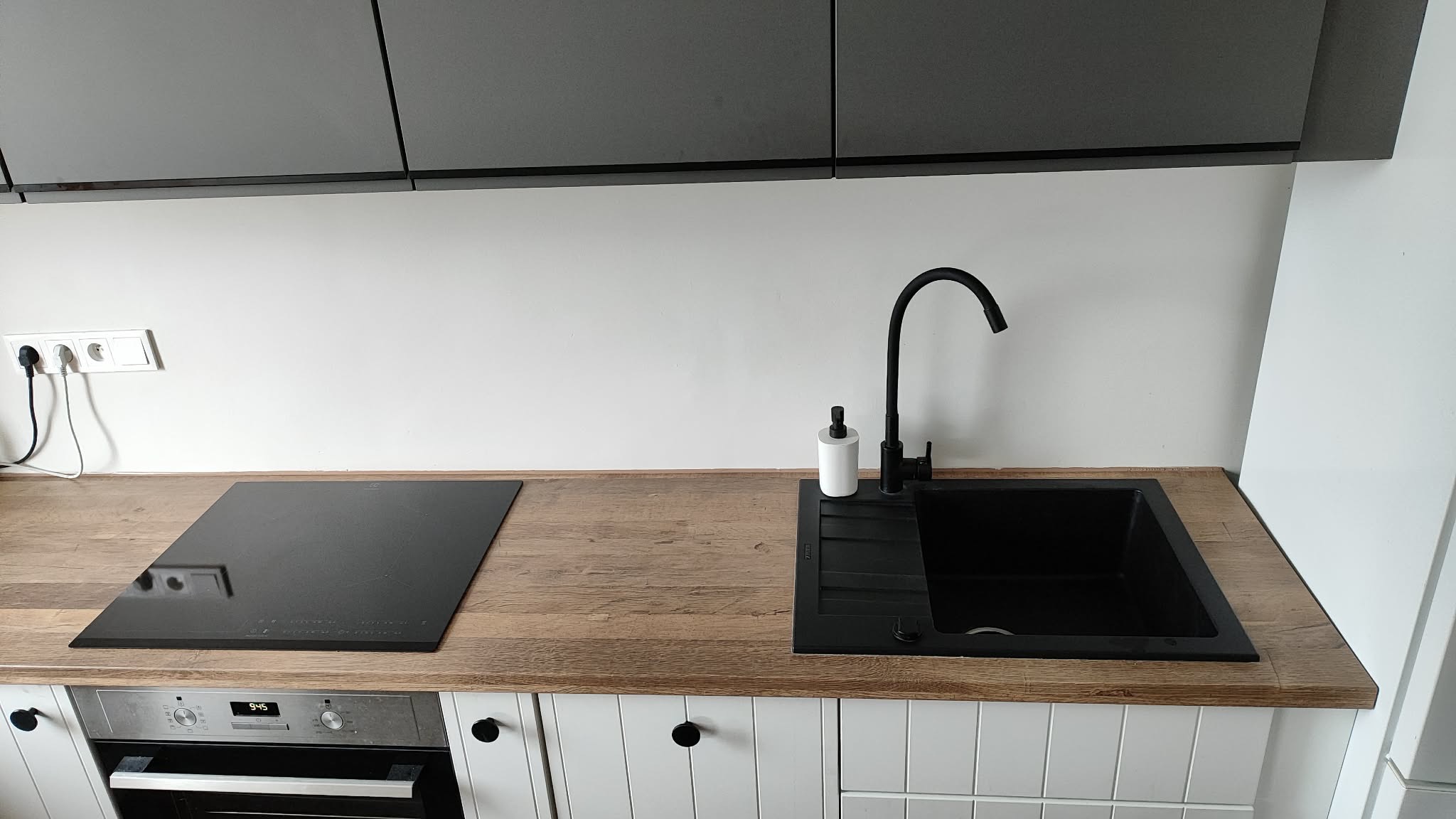 Nowoczesna kuchnia po remoncie: czarny zlew i płyta indukcyjna na drewnianym blacie, białe szafki z czarnymi uchwytami, szare szafki wiszące, minimalistyczny design.