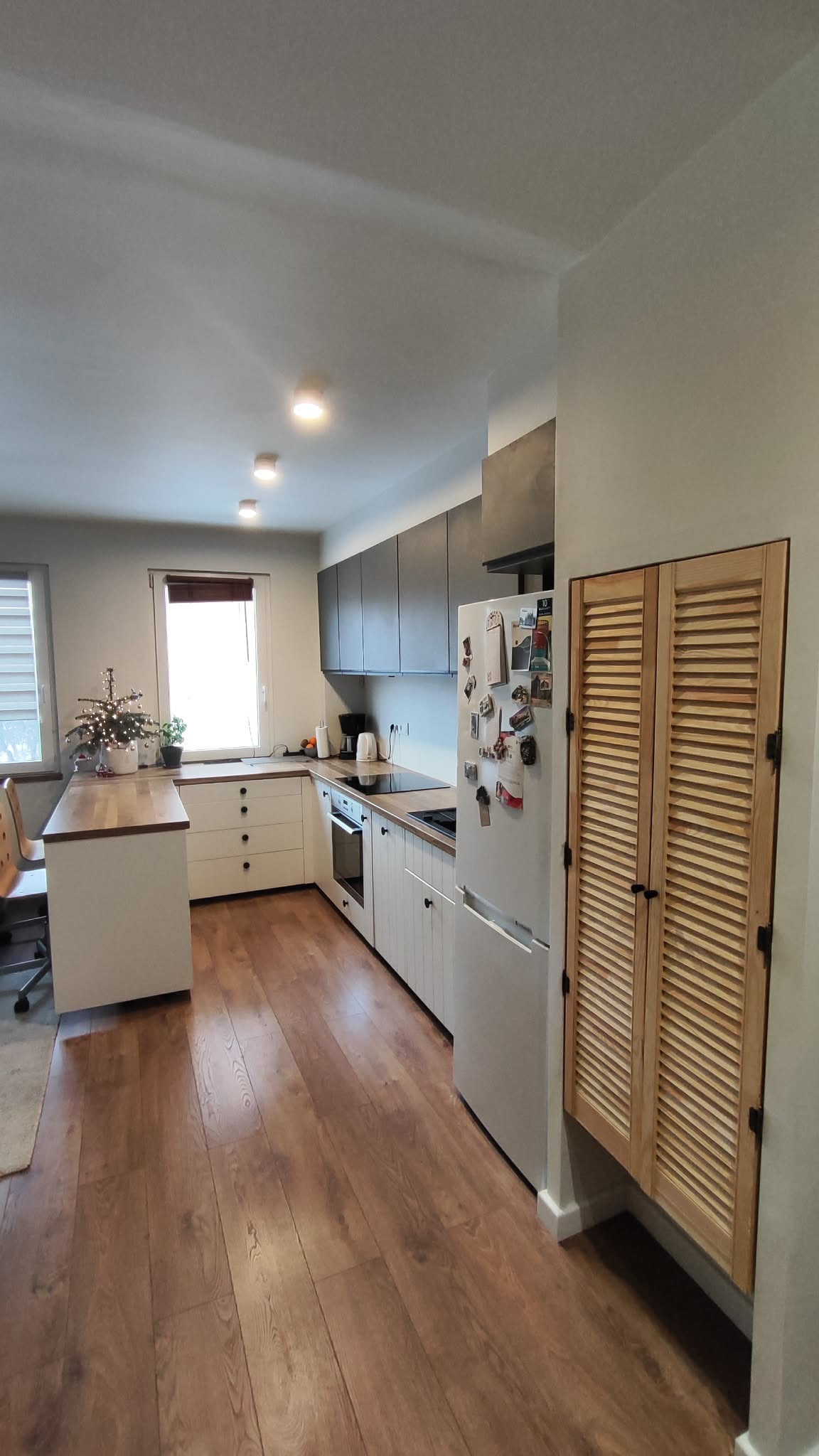 Nowoczesna kuchnia z drewnianą podłogą, białymi szafkami i szarymi górnymi szafkami. Na blacie stoi mała choinka, a na lodówce wiszą magnesy.