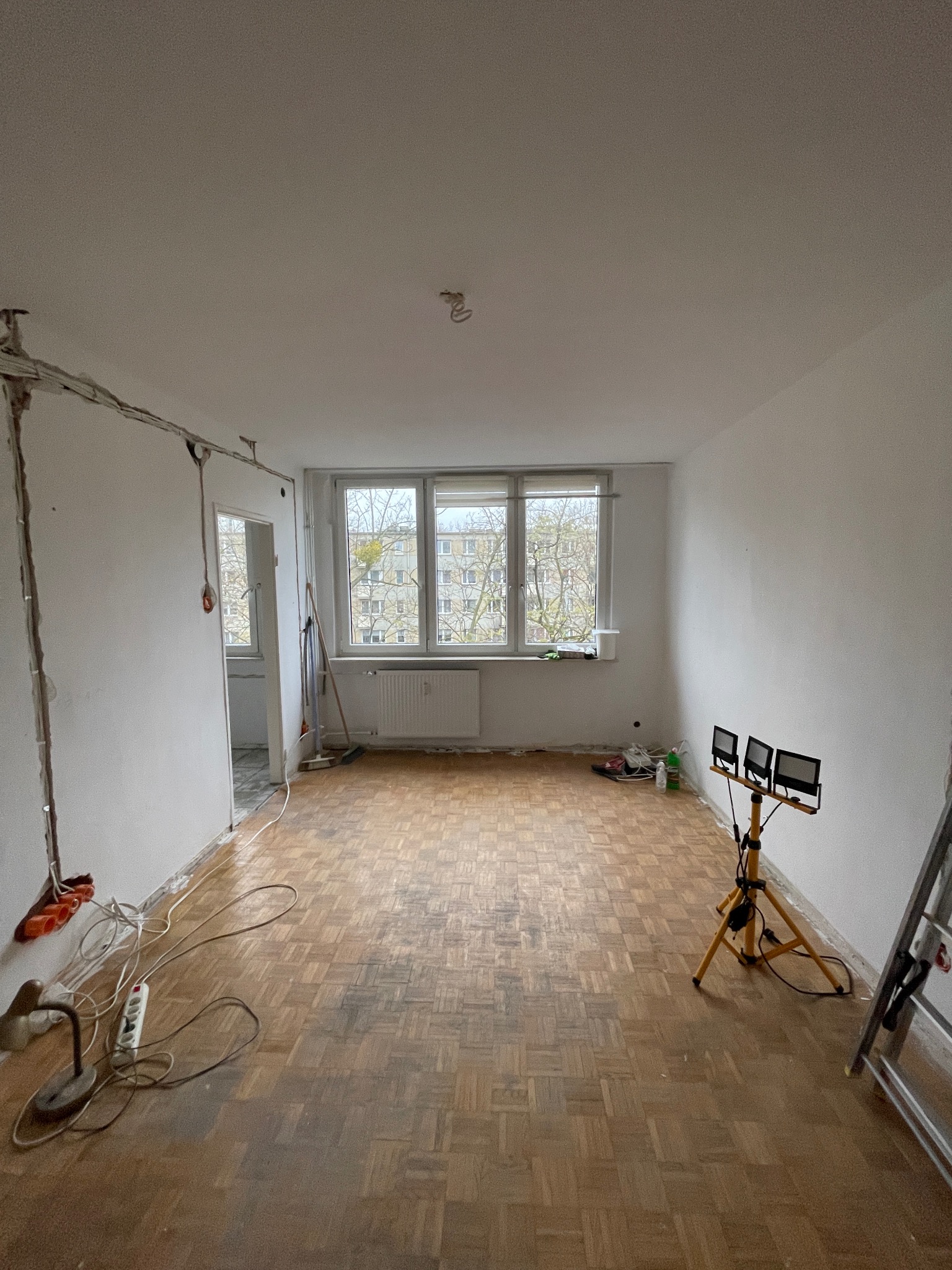 Pokój w trakcie remontu: odsłonięte przewody elektryczne, drewniana podłoga, drabina, lampa budowlana. Widok na okno z drzewami. Przygotowanie do prac wykończeniowych.