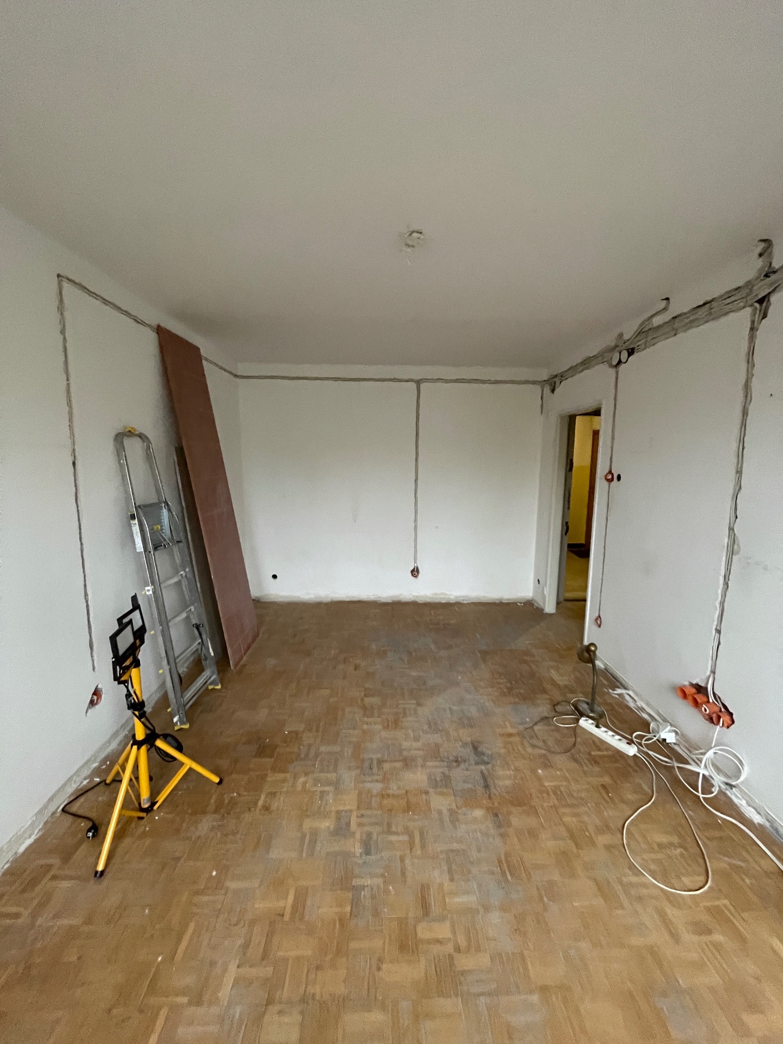 Pokój w trakcie remontu: widoczne kable elektryczne w ścianach, drabina, lampa budowlana i parkiet. Przygotowanie do prac wykończeniowych.