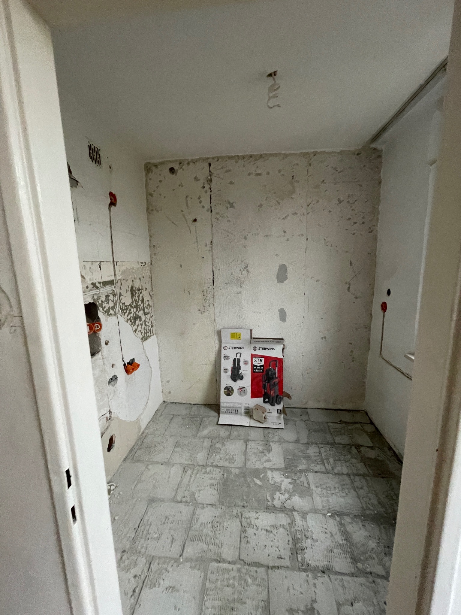 Wnętrze łazienki w trakcie demontażu płytek, widoczne ślady po kleju na ścianach i podłodze. Na podłodze stoją kartony z nowym sprzętem.