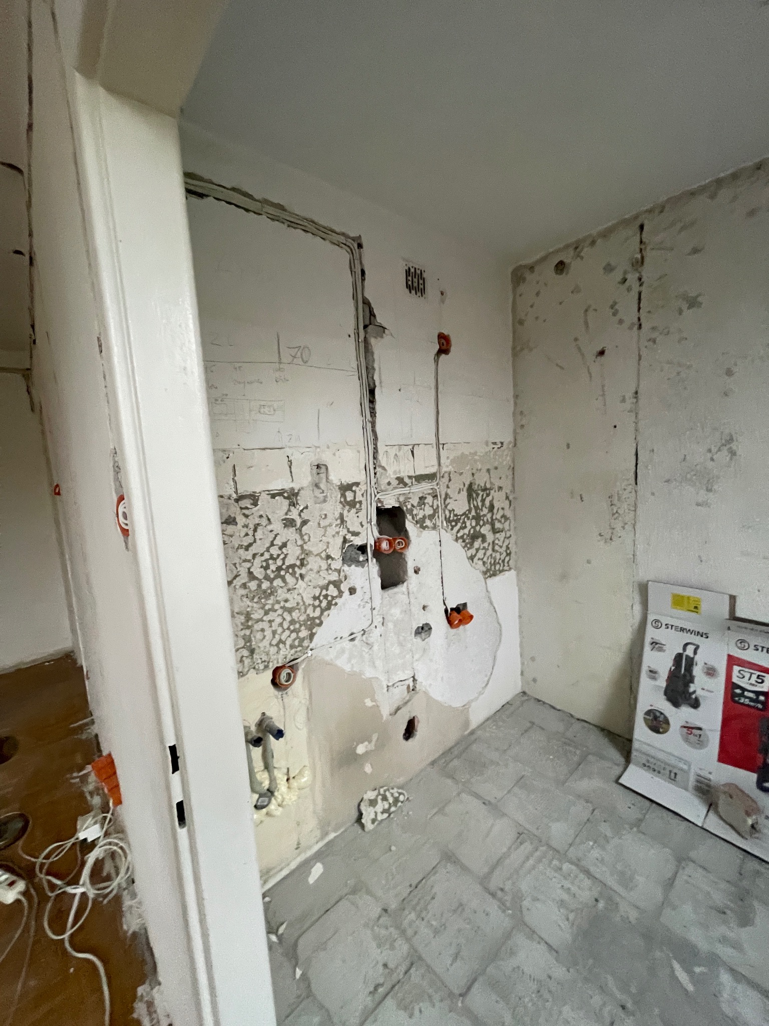 Wnętrze w trakcie demontażu instalacji w pomieszczeniu. Odsłonięte przewody elektryczne i rury wodne na ścianie. Widoczne ślady po starych płytkach i instalacjach.