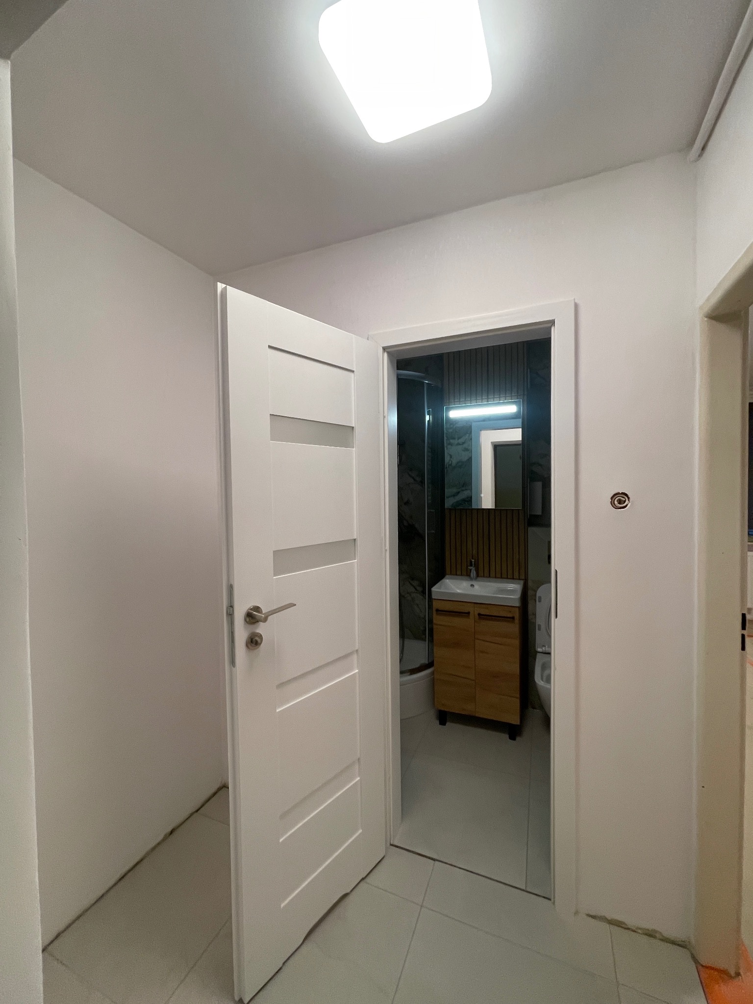 Otwarta biała łazienkowa z drewnianą szafką pod umywalką i kabiną prysznicową, widoczna z korytarza. Minimalistyczny design, jasne oświetlenie.