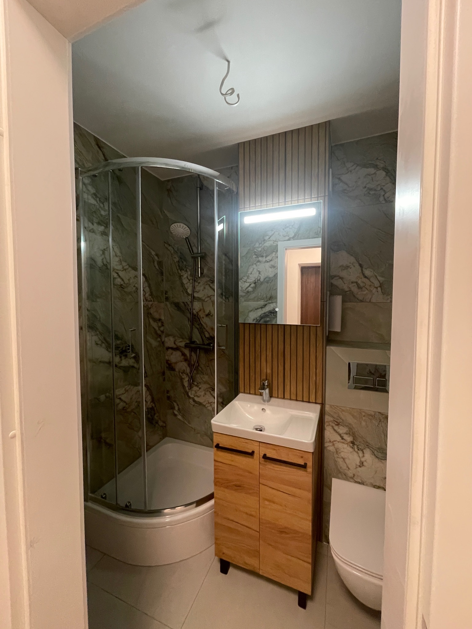 Nowoczesna łazienka z kabiną prysznicową, umywalką na szafce z drewnianym wzorem i toaletą. Ściany wyłożone płytkami imitującymi marmur. Oświetlenie LED nad lustrem.