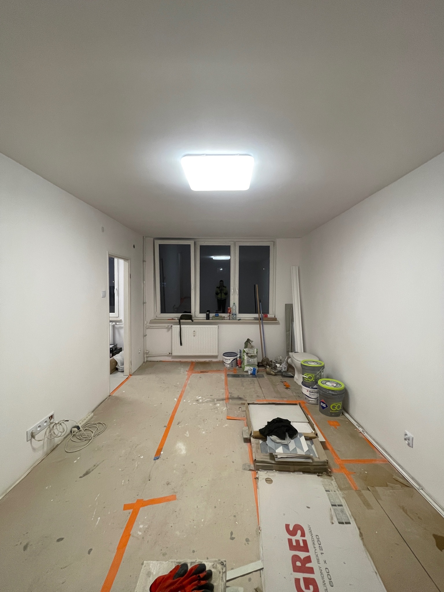 Pokój w trakcie remontu: widoczne płytki GRES, farby, narzędzia, pomarańczowe linie wyznaczające obszar roboczy. W oknie odbija się sylwetka osoby.