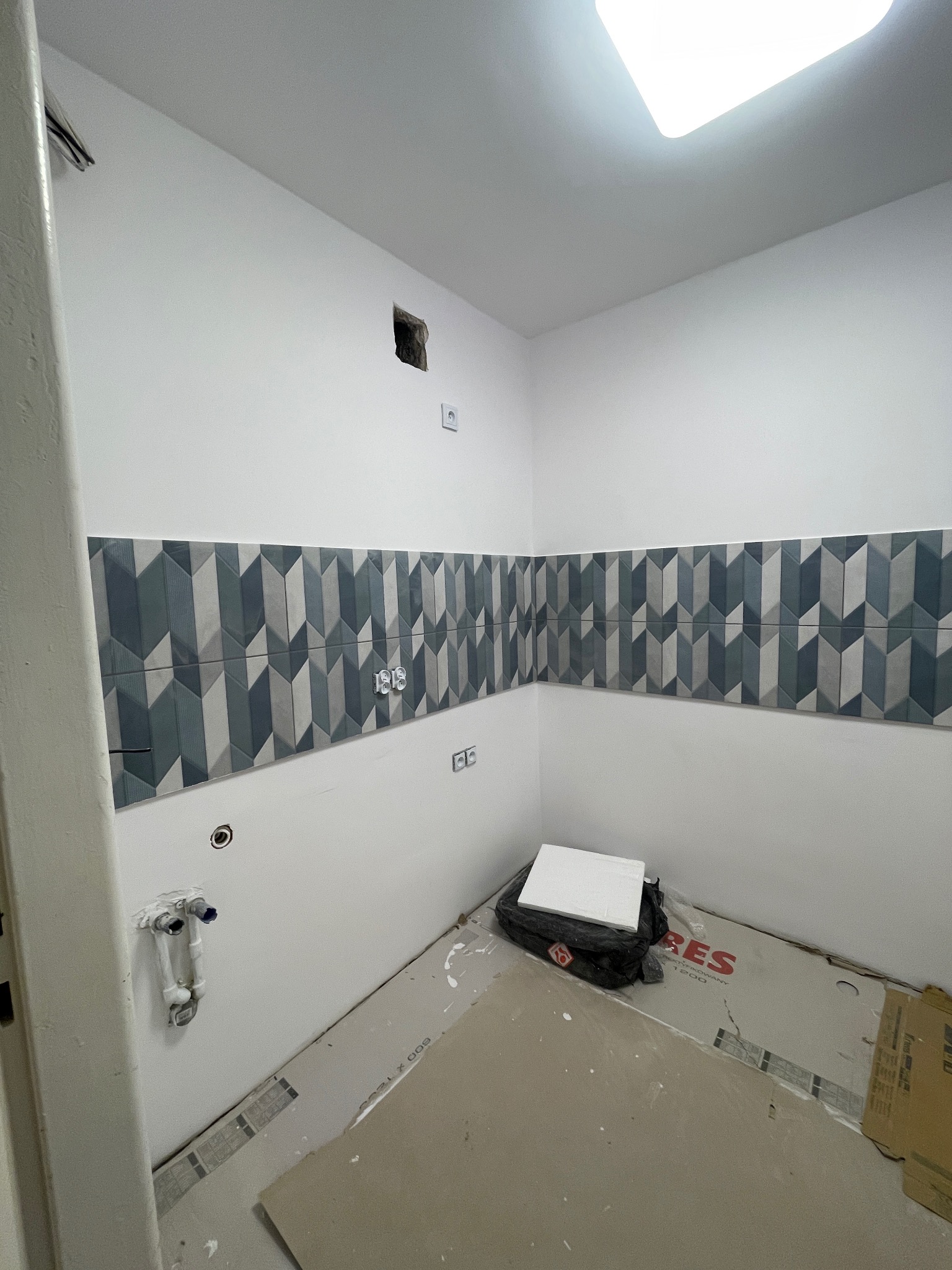 Układanie dekoracyjnych płytek w geometryczny wzór w łazience w Warszawie. Widoczne przygotowane płytki i instalacje. Świeżo położona glazura na ścianie.