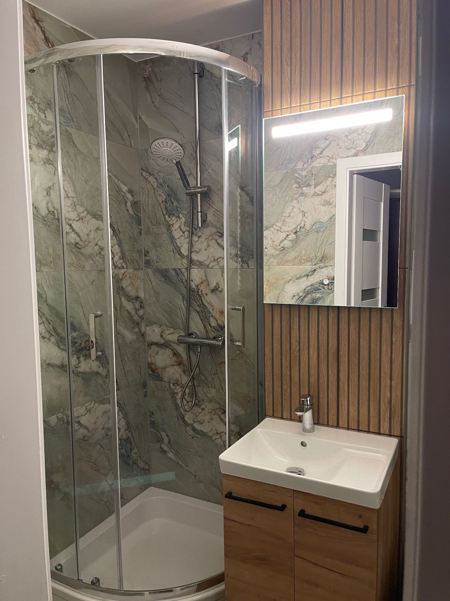 Wykończona łazienka z kabiną prysznicową, umywalką z szafką w odcieniach drewna i lustrem z oświetleniem. Marmurowe płytki na ścianach i drewniane panele dekoracyjne.