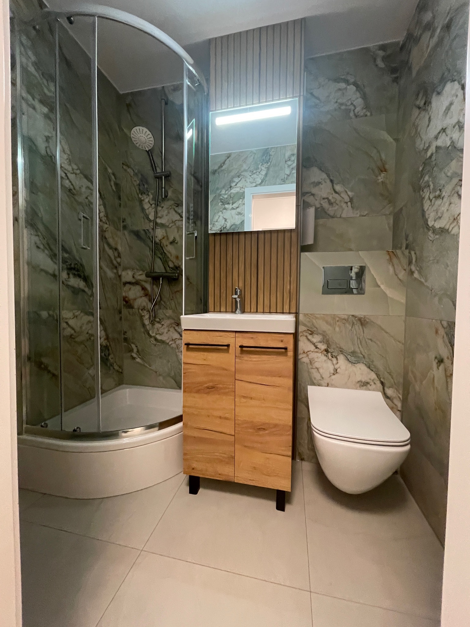 Nowoczesna łazienka z kabiną prysznicową, umywalką z szafką z drewna i podwieszaną toaletą. Marmurowe płytki na ścianach i jasna podłoga.