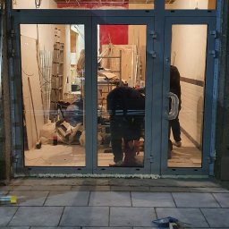 DG SPÓŁKA Z OGRANICZONĄ ODPOWIEDZIALNOŚCIĄ - Montaż szklanych drzwi wejściowych z szarymi ramami. W tle widoczne wnętrze w trakcie remontu: drabina, materiały budowlane, instalacja sufitowa.